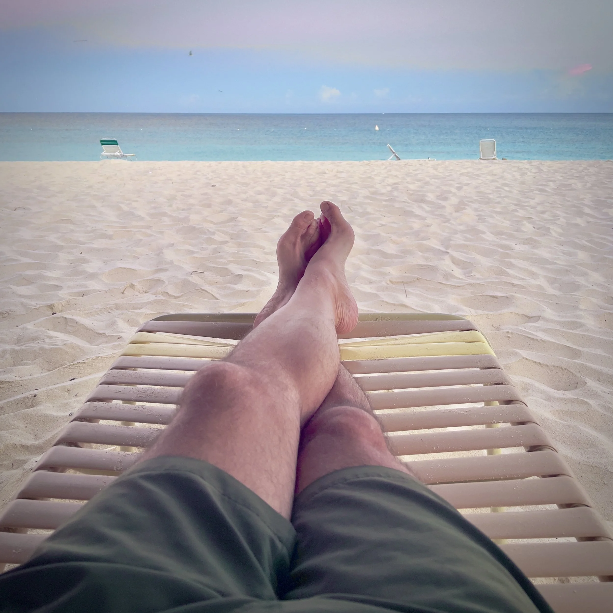 Person relaxing on a lounge chair at the beach, with legs crossed, overlooking the sandy beach, ocean, and clear sky.