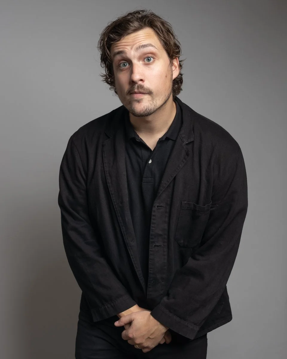 A man with light skin, curly brown hair, and a beard, looking at the camera with a surprised expression, wearing a black jacket and black shirt against a gray background.