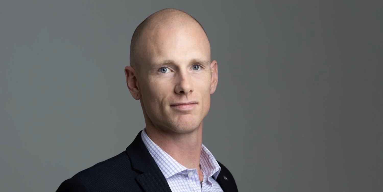 A portrait of a bald man with light skin, blue eyes, wearing a dark suit jacket and a checkered collared shirt against a plain gray background.