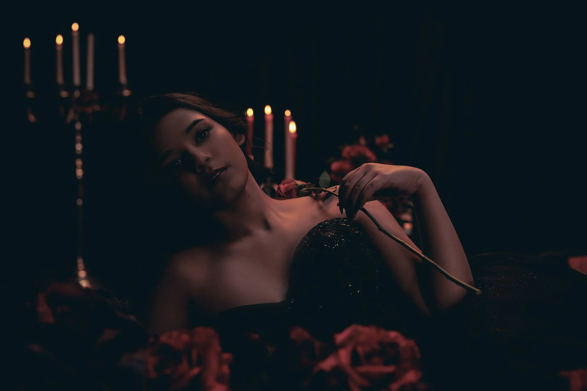 A woman lying in a dark room, holding a pink rose, with lit candles in the background, creating an intimate, dramatic ambiance.