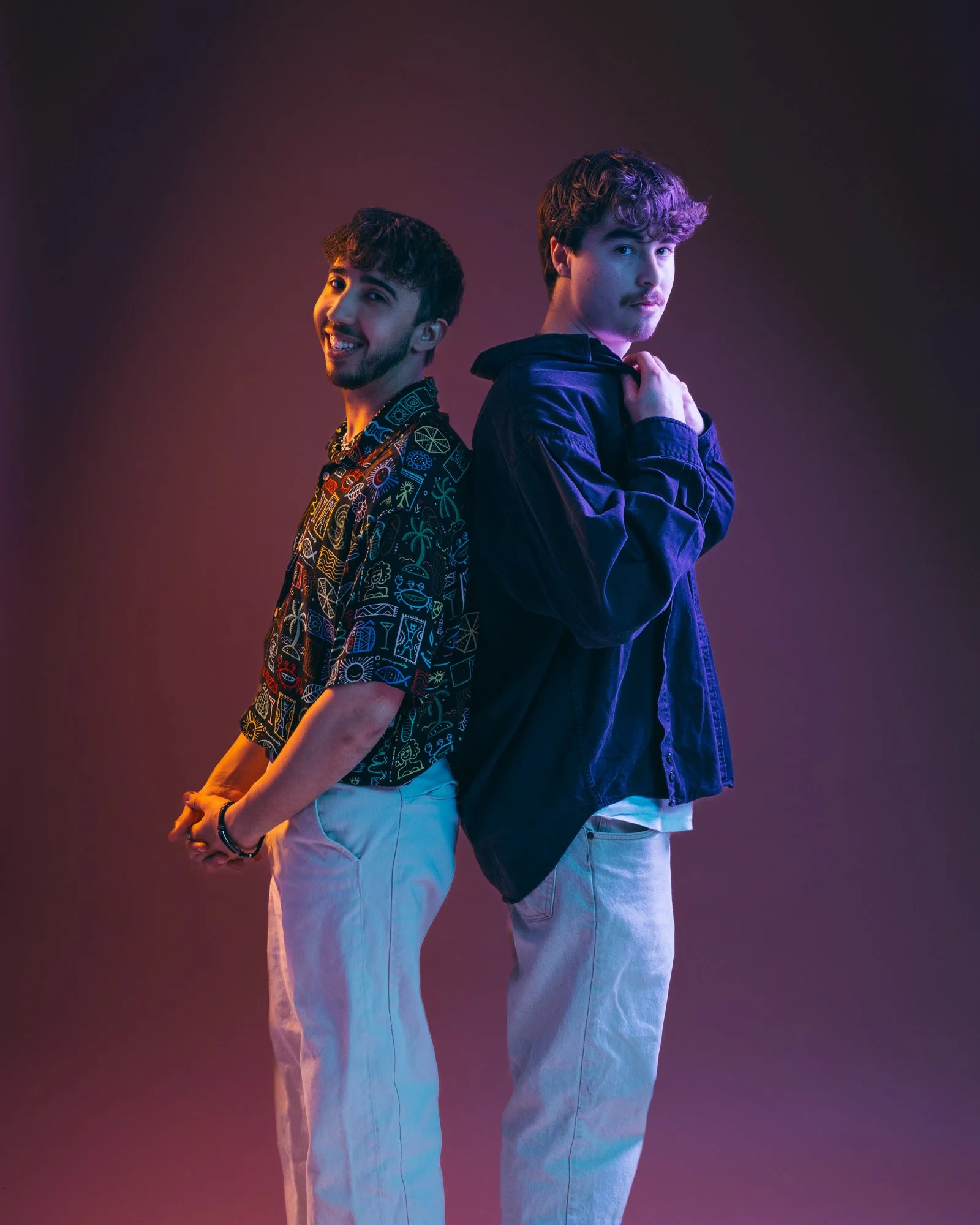 Two young men standing back to back, one smiling and the other with a serious expression, in a colorful studio with purple and blue lighting.