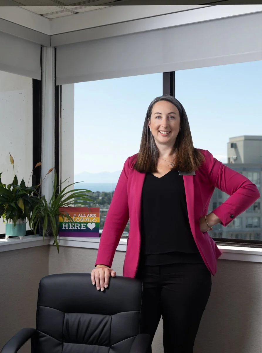 A woman in a pink blazer standing in an office near a window with a view of city buildings and water. She is smiling, holding a black office chair, with plants and a colorful sign that says 'All Are Welcome Here' on the windowsill.