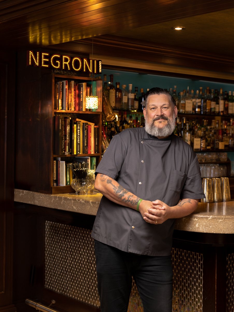A bearded man in a gray chef's coat with tattoos on his arms standing behind a bar counter, smiling, with bottles on shelves and books nearby, in a warmly lit bar or restaurant setting.