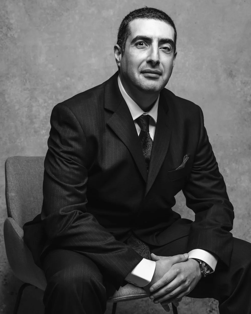 Black and white portrait of a man in a suit sitting on a chair against a plain background, looking at the camera with a confident expression.