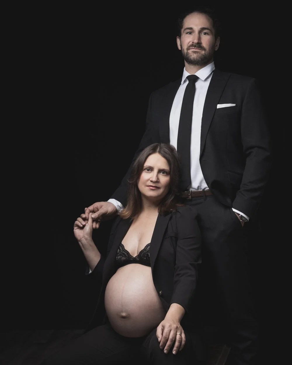 A pregnant woman in a black blazer and lace bra sits in front while a man in a black suit and tie stands behind her with his hand on her shoulder, both against a black background.