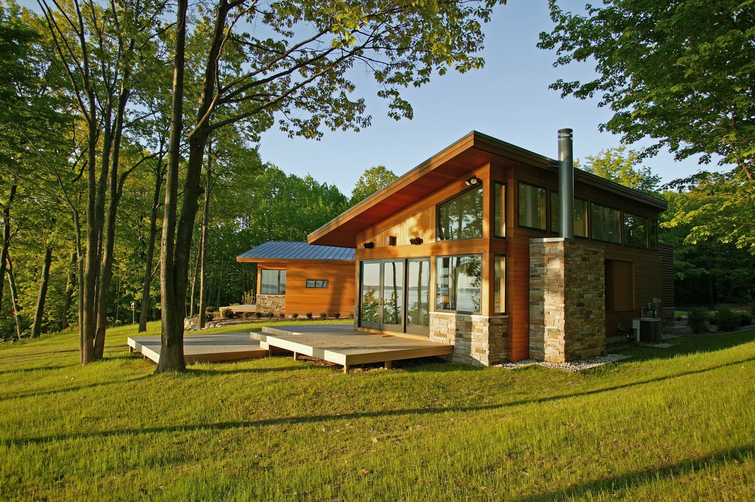 CARLYON RESIDENCE . Traverse City, MI This 3,000 square foot residence is set within two acres bound by dense trees, but open to a view of Grand Traverse Bay on Lake Michigan. The landscape and view directly informed the geometry and spatial organization of the home. From the approach through a thick grove of trees to an open sunlit yard, to movement through the interior spaces, the residence is experienced as an orchestrated procession of views. The intent was that view and space is slowly revealed as one moves deeper into the site, and then further into the home itself. The west façade one views on approach, is intended to be a ‘formal front’, a collage of solid and opaque materials layered together to express separations in program, but revealing little of what is inside. The horizontal batten cedar cladding is cut open and lifted to become a louvered sunscreen for the double height entry space, which glows like a lantern in the evening. Utilitarian spaces such as garage, laundry, and bathrooms occupy this façade, granting prominence to more important functions to take advantage of the view. This gives the home a compelling duality that works both environmentally and programmatically. The mostly solid and louvered southwest exposures protect against summer sun, whilst the open and glassy east façade welcomes daylight in the morning and provides breathtaking views from the living and master bedroom spaces. The double height entry space becomes the fulcrum, uniting two equally important, but programmatically different public and private wings of the home. Boundaries of inside and outside are blurred. From the entry, green slate paving continues out to a patio, becoming an outdoor room open only to the bay view beyond. Cherry flooring continues out to cedar deck, and clerestory windows invite views of the tree canopy above. As one travels through the home, every movement is terminated with a view of the outside, extending the perceived spatial boundaries. All photos © Glen Rauth Photography except slides 1,4,5 and 6 by STUDIOrobert jamieson