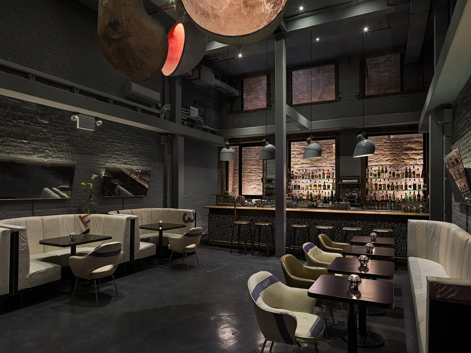 The Ship . New York, NY The client for this project came to me with a site and a name, The Ship . What ensued was a dramatic spatial change that took a long, low ceiling ground floor space with a dark basement, into a hip industrial bar with high ceilings. Occupancy and egress requirements for the space limited the allowable ‘public’ area within the bar. Instead of having excess back of house, we removed about a third of the ground floor. The subtly marked entry at the street leads to a non-descript long, low corridor at the end of which unfolds a dramatic double height space that one descends down to. The bar’s name is alluded to in the materiality and design, but in a modern way, with riveted aged zinc wall panels, custom booths upholstered with reclaimed patchwork sailcloth, and a dynamic lighting installation of inverted antique ship ventilation shafts. The bar utilizes many other found objects such as the common washbasin, antique doors and custom fabricated mirrors in the bathroom. Both the stair treads and window frames were made from the floor joists that were removed to open up the space. All photos © Sam Oberter Photography