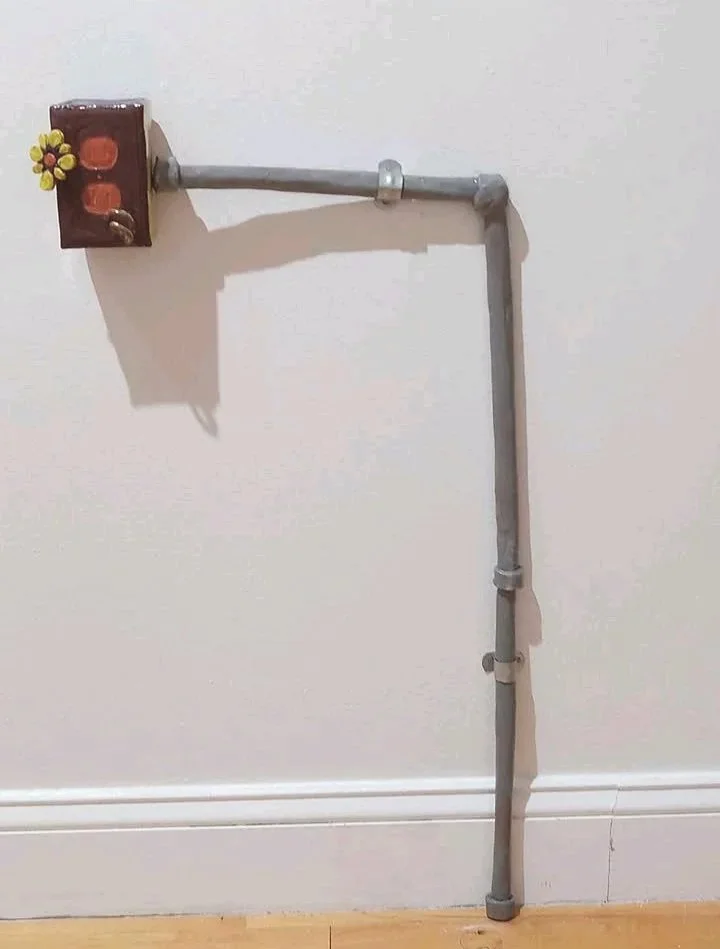 A DIY wall-mounted pipe fixture with a small wooden electrical box attached, decorated with a flower ornament, against a plain white wall.