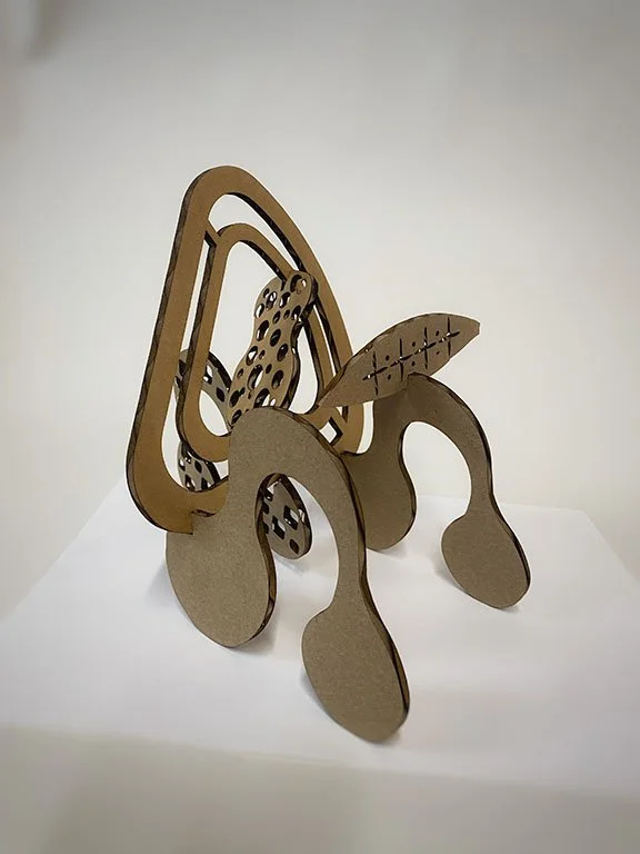 A cardboard sculpture resembling a whimsical animal, possibly a dog or a creature, made of layered, cut-out cardboard pieces with various shapes and patterns.