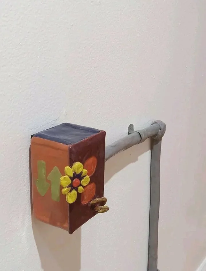 Colorful handmade art piece attached to a wall pipe, featuring a flower design and the word 'love' painted on it.
