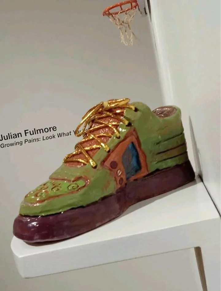 Ceramic sculpture of a colorful sneaker with gold laces, placed on a white surface. In the background, part of a small basketball hoop is visible.