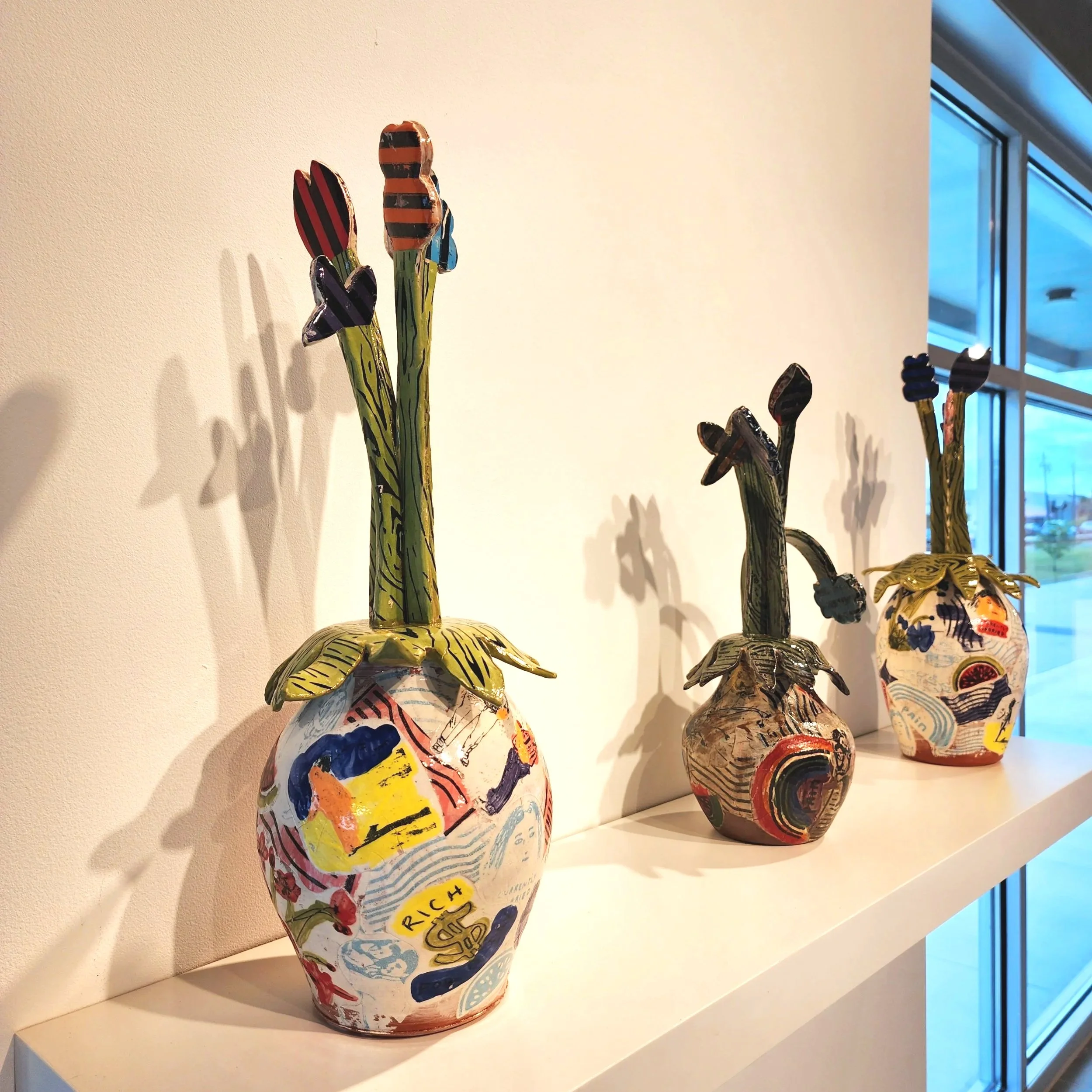 Three colorful ceramic vases with artistic designs on a white shelf near a window.