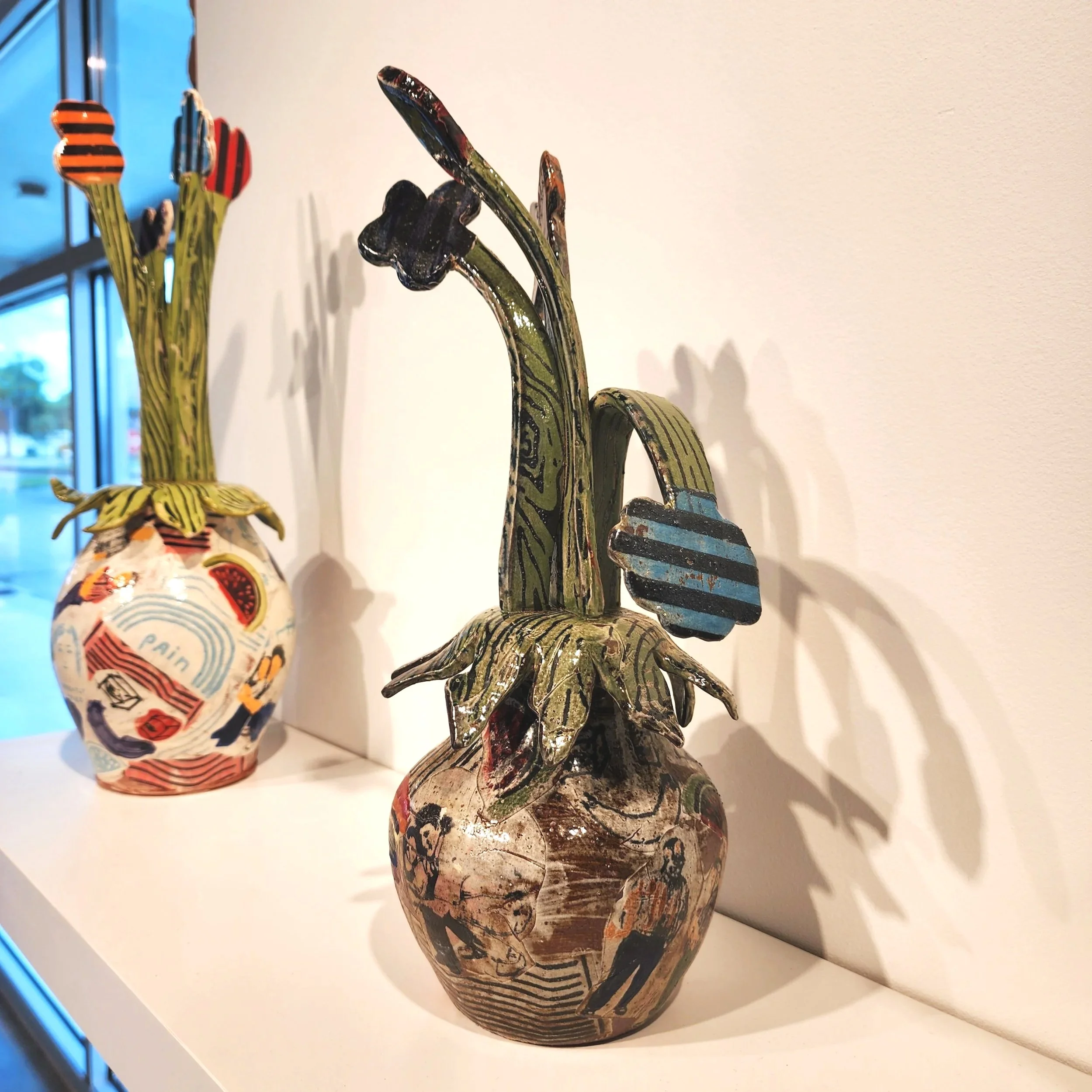 Decorative ceramic vases with painted images and patterns, shaped like flowers, placed on a white surface near a window.