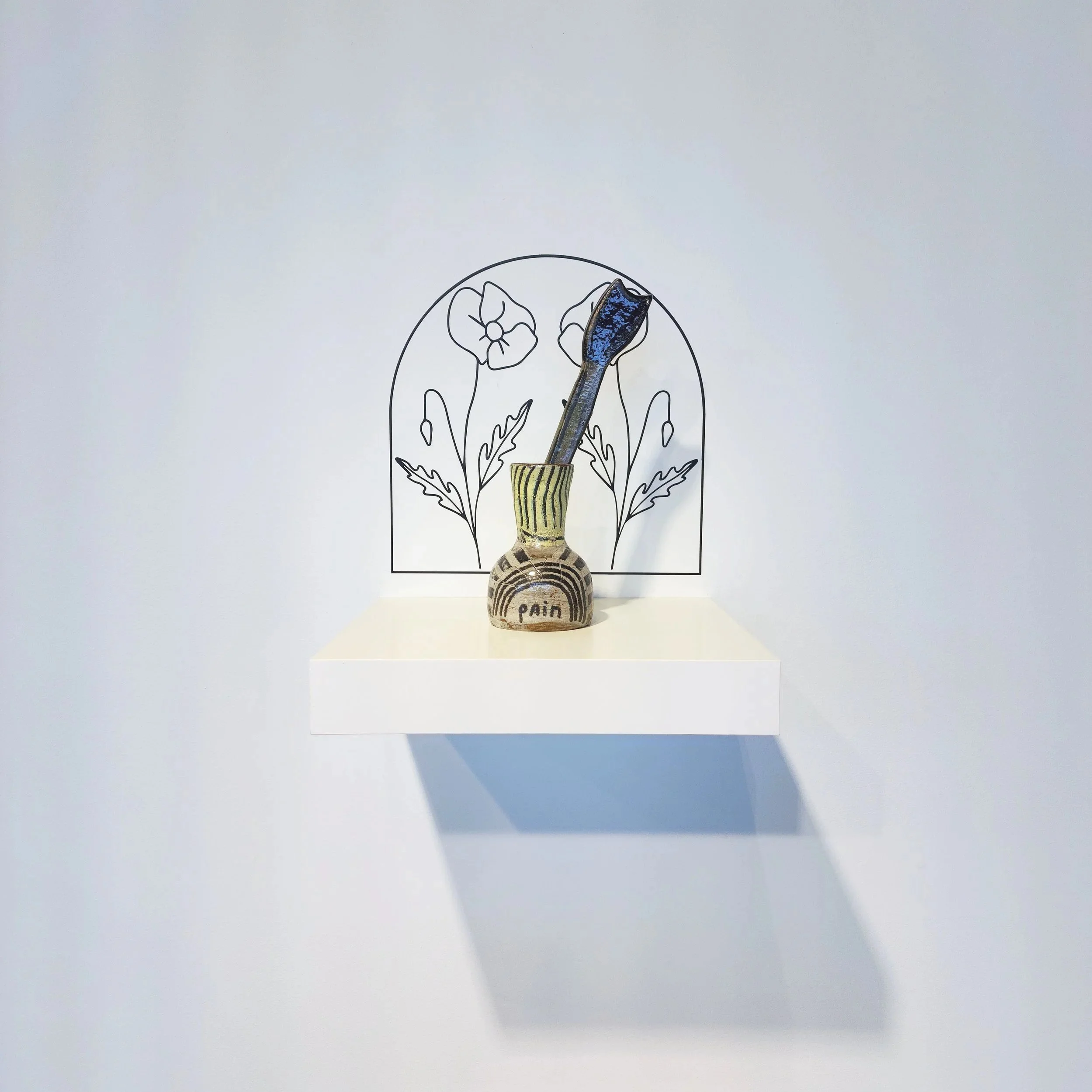 Ceramic vase labeled 'pain' with a clay flower inside, placed on a white shelf against a wall. The wall features a simple black line drawing of flowers as decoration.