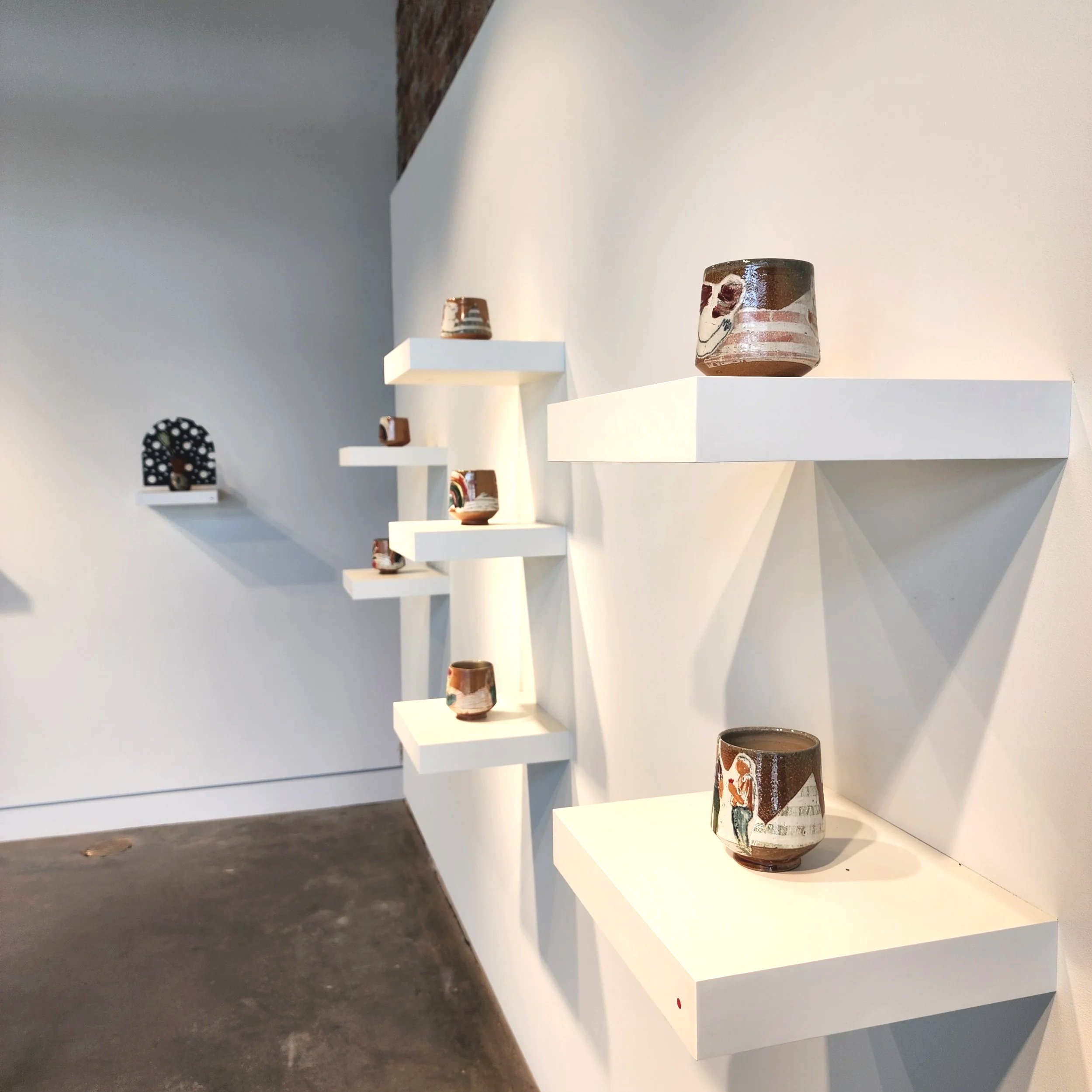 Pottery displayed on white floating shelves against a white wall in an art gallery.