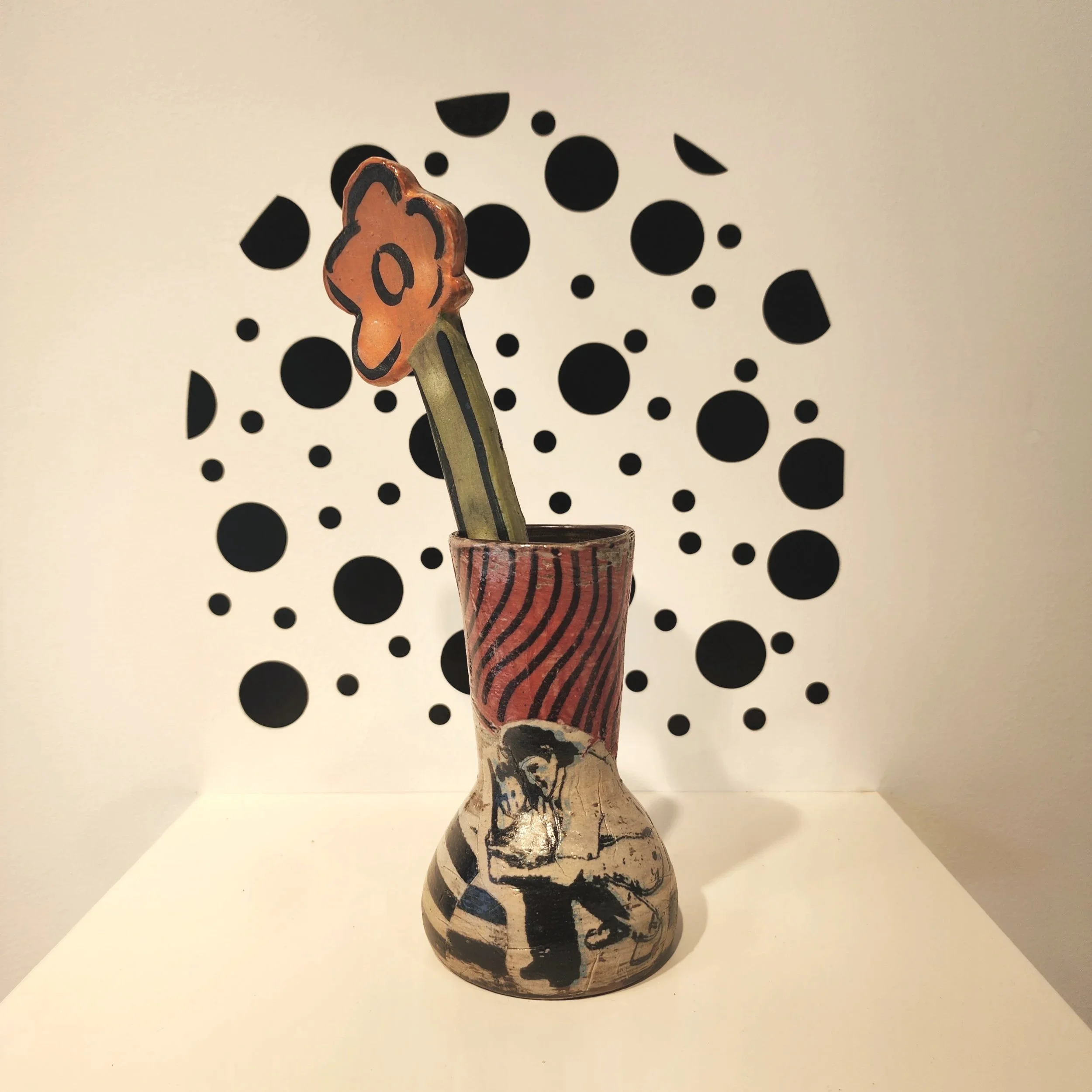 Decorative vase with a painted illustration of a person, containing a whimsical flower with a face, set against a backdrop of black polka dots.