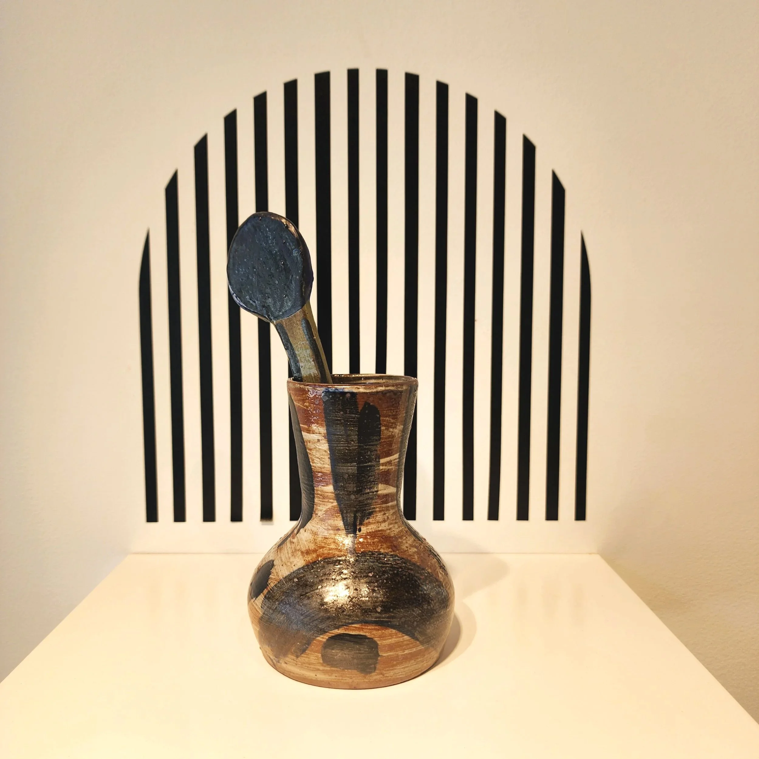 A ceramic vase with a clay flower inside, set against a backdrop with black vertical stripes.
