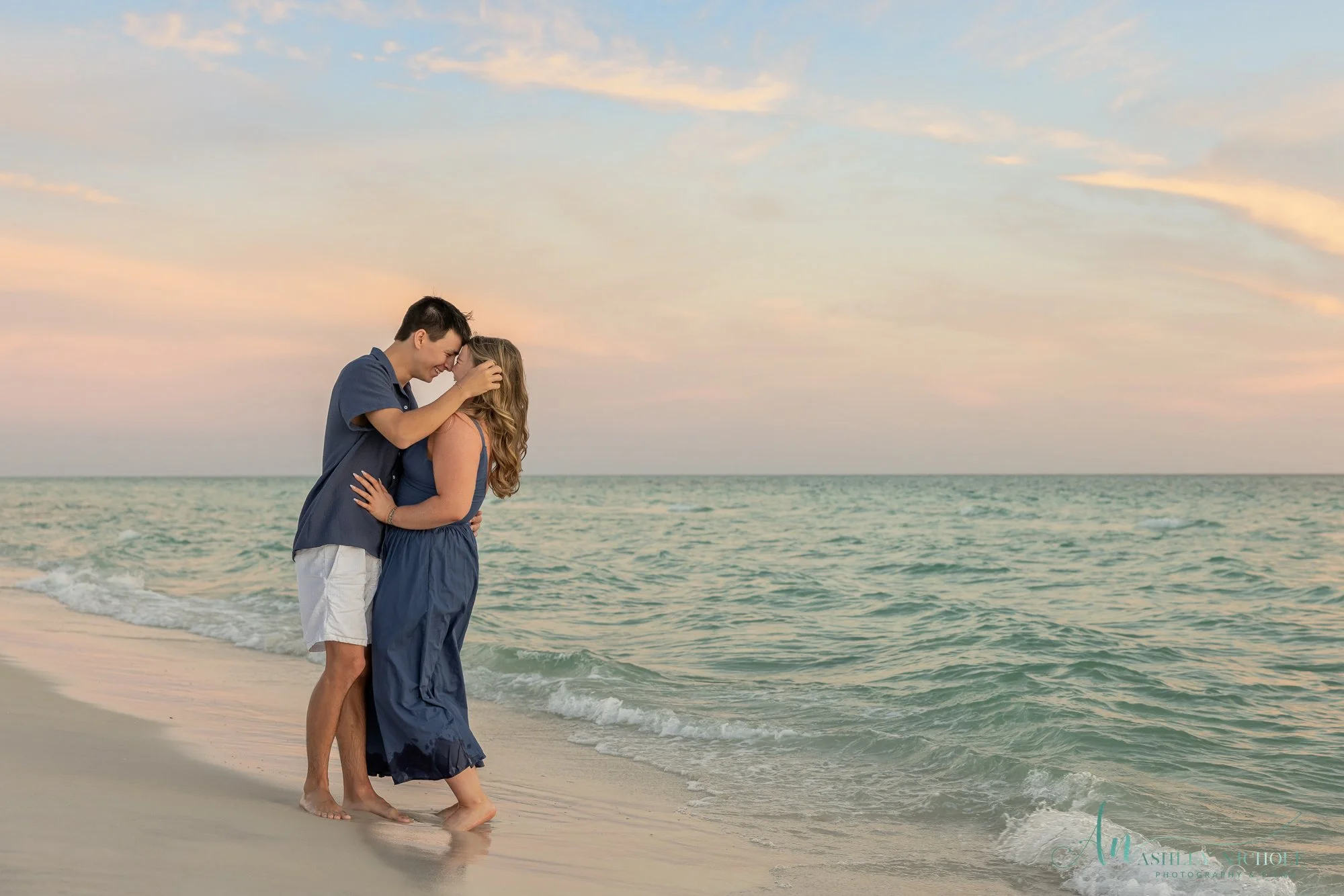 A couple standing on the beach by the ocean, touching foreheads and smiling at each other during sunset.