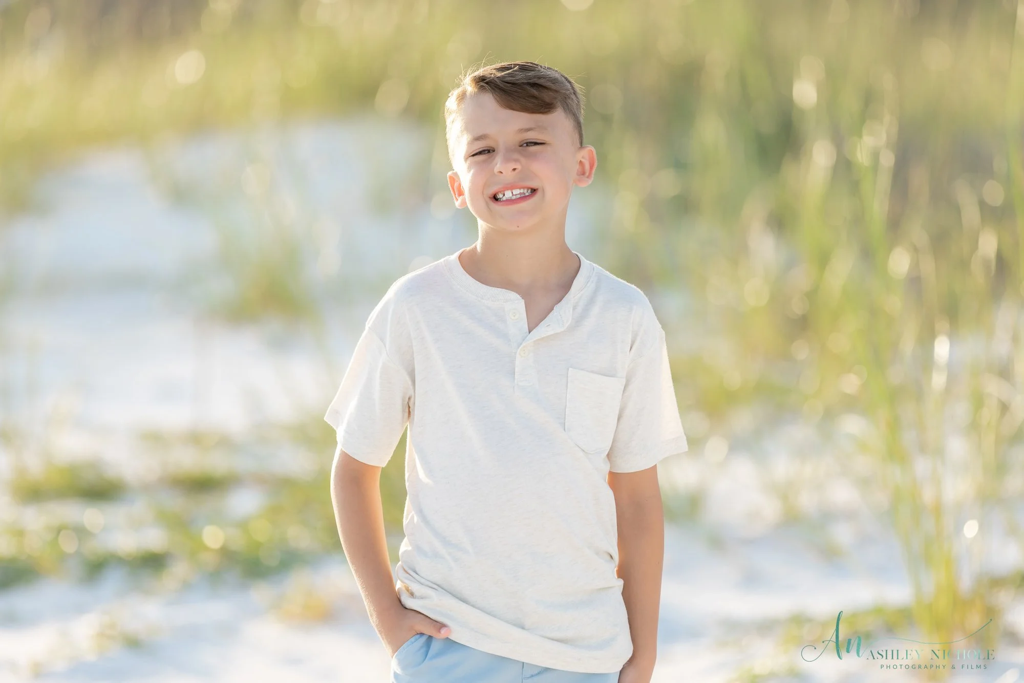 Young boy smiling outdoors near water with tall grass in the background.