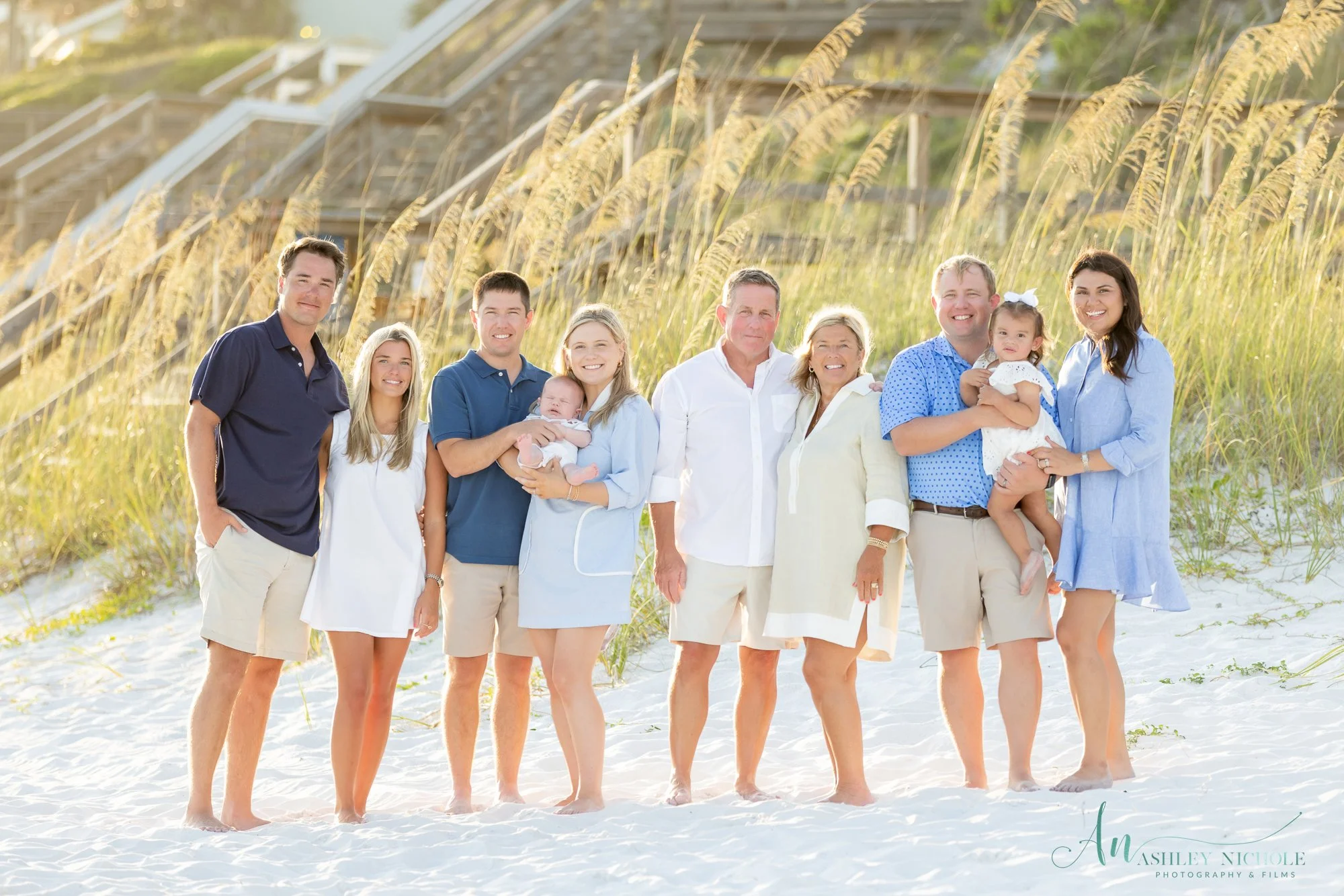 A multi-generational family poses on a sandy beach in front of dunes and tall grass, all dressed in casual summer clothing, smiling at the camera.