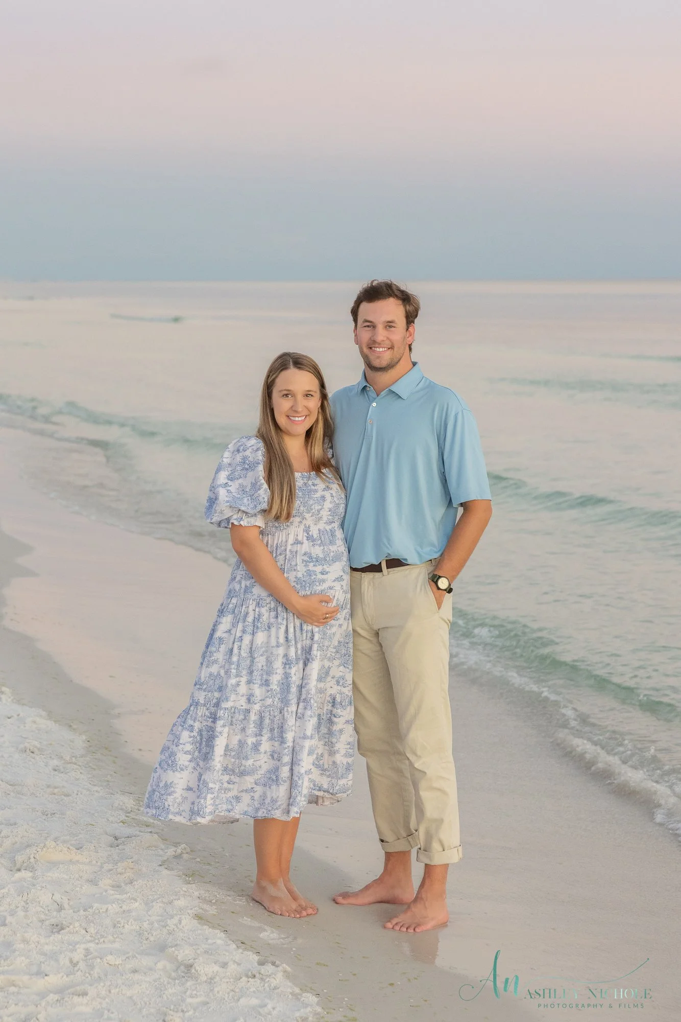 A couple standing barefoot on the beach near the shoreline with gentle waves and a colorful sky in the background.