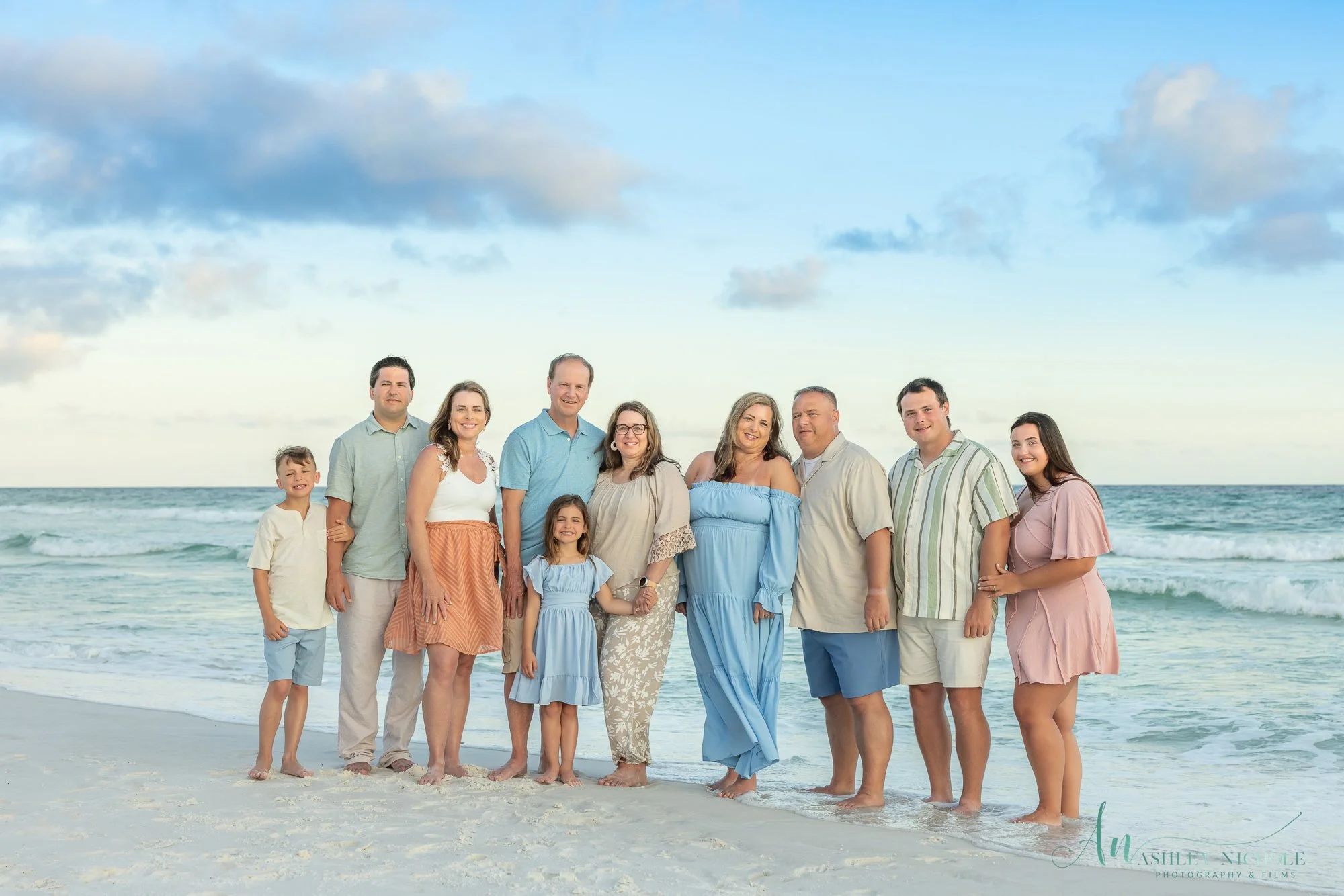 A family of ten standing on a sandy beach near the ocean with waves and a partly cloudy sky in the background.