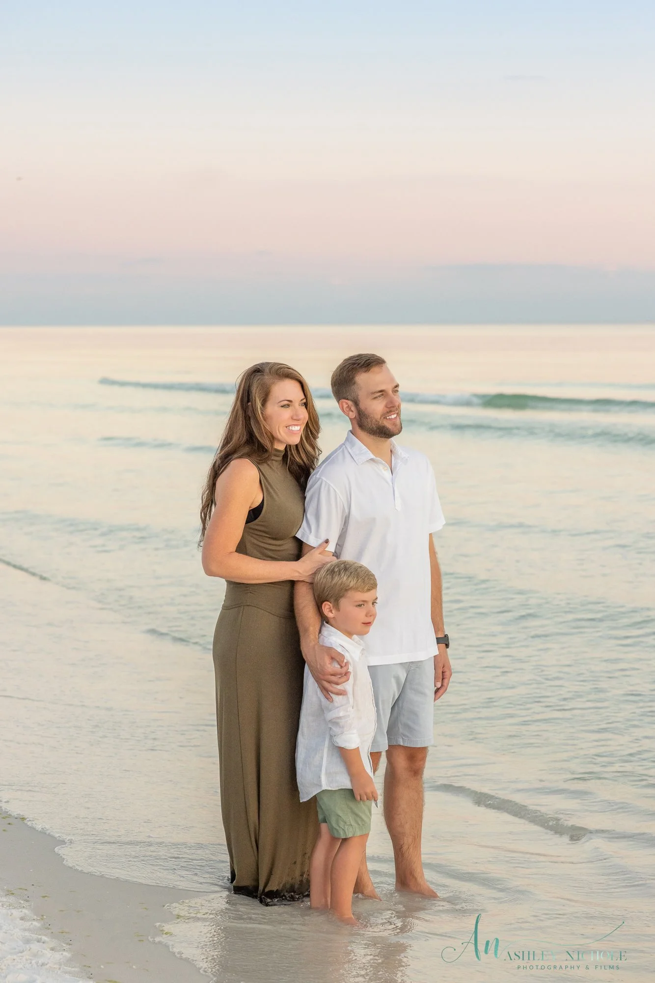 A family standing on the beach near the water at sunset, smiling and looking out towards the ocean.