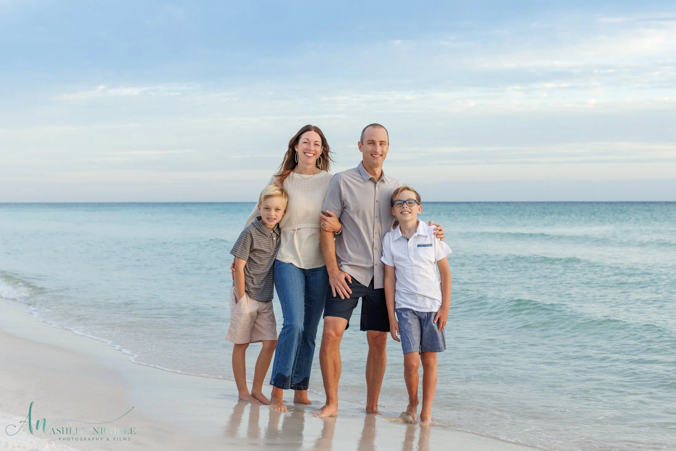 A family of four standing on a sandy beach near the ocean with their feet in the water, smiling and holding each other during a clear day with a partly cloudy sky.