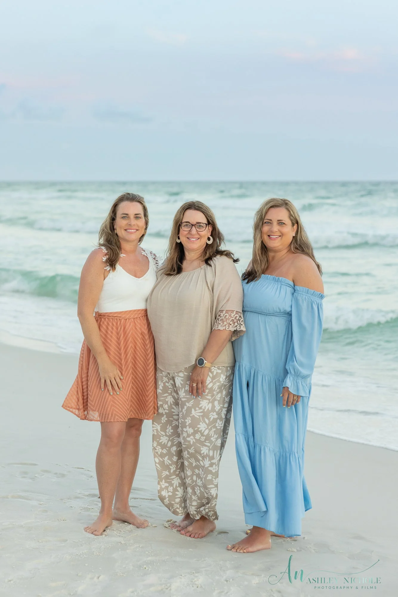 Three women standing barefoot on the beach with ocean waves in the background, smiling at the camera, during daytime.