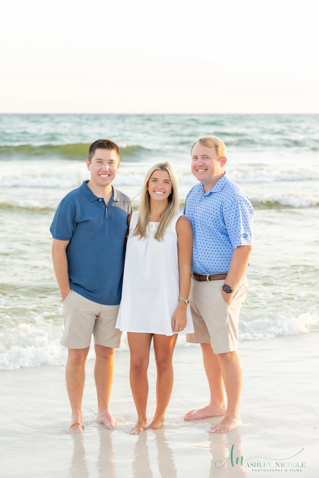 Three people standing on the beach near the water, smiling at the camera, with the ocean waves behind them.