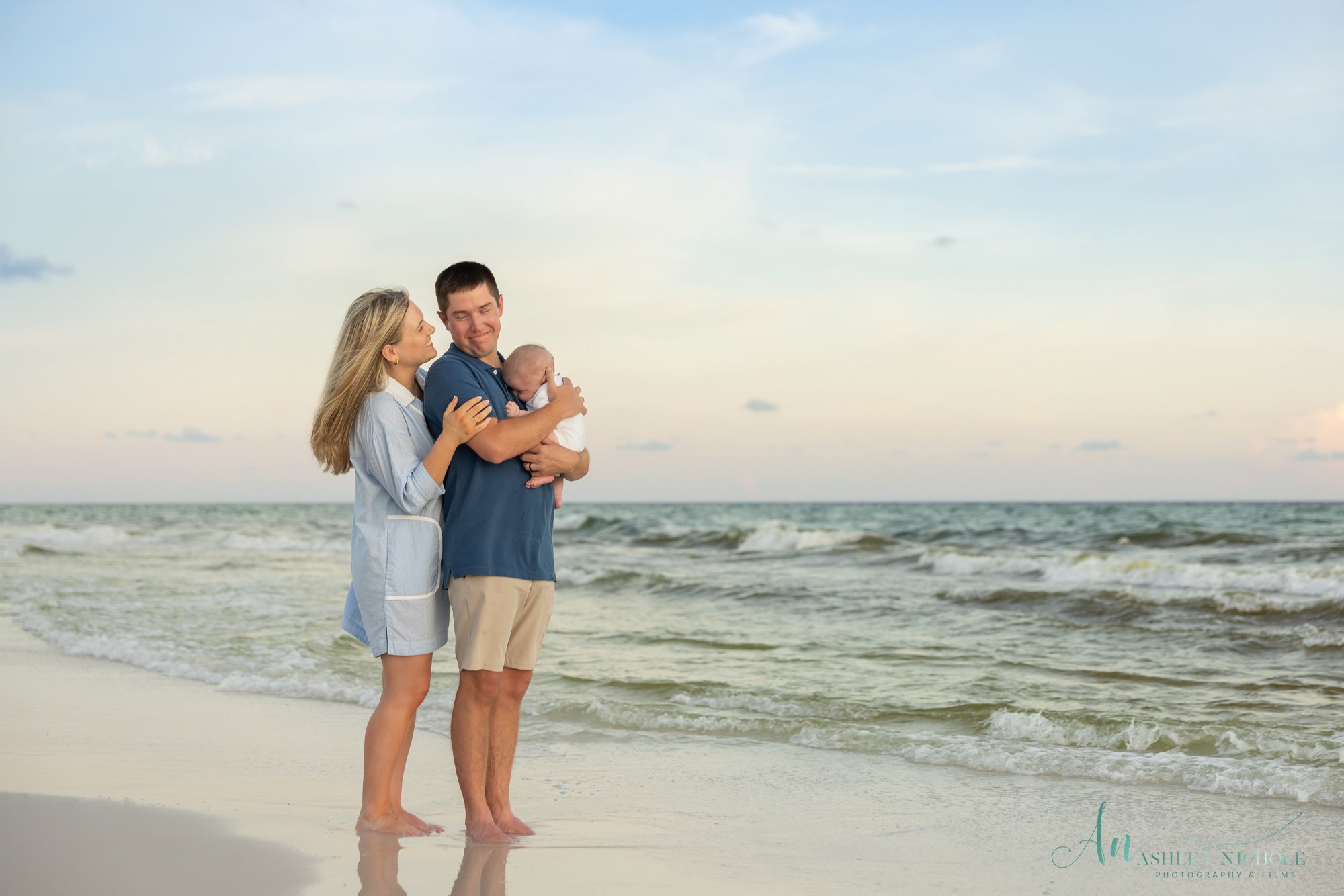 A family of three, a mother, father, and a baby, standing on the beach near the ocean at sunset, with the mother and father embracing the baby.