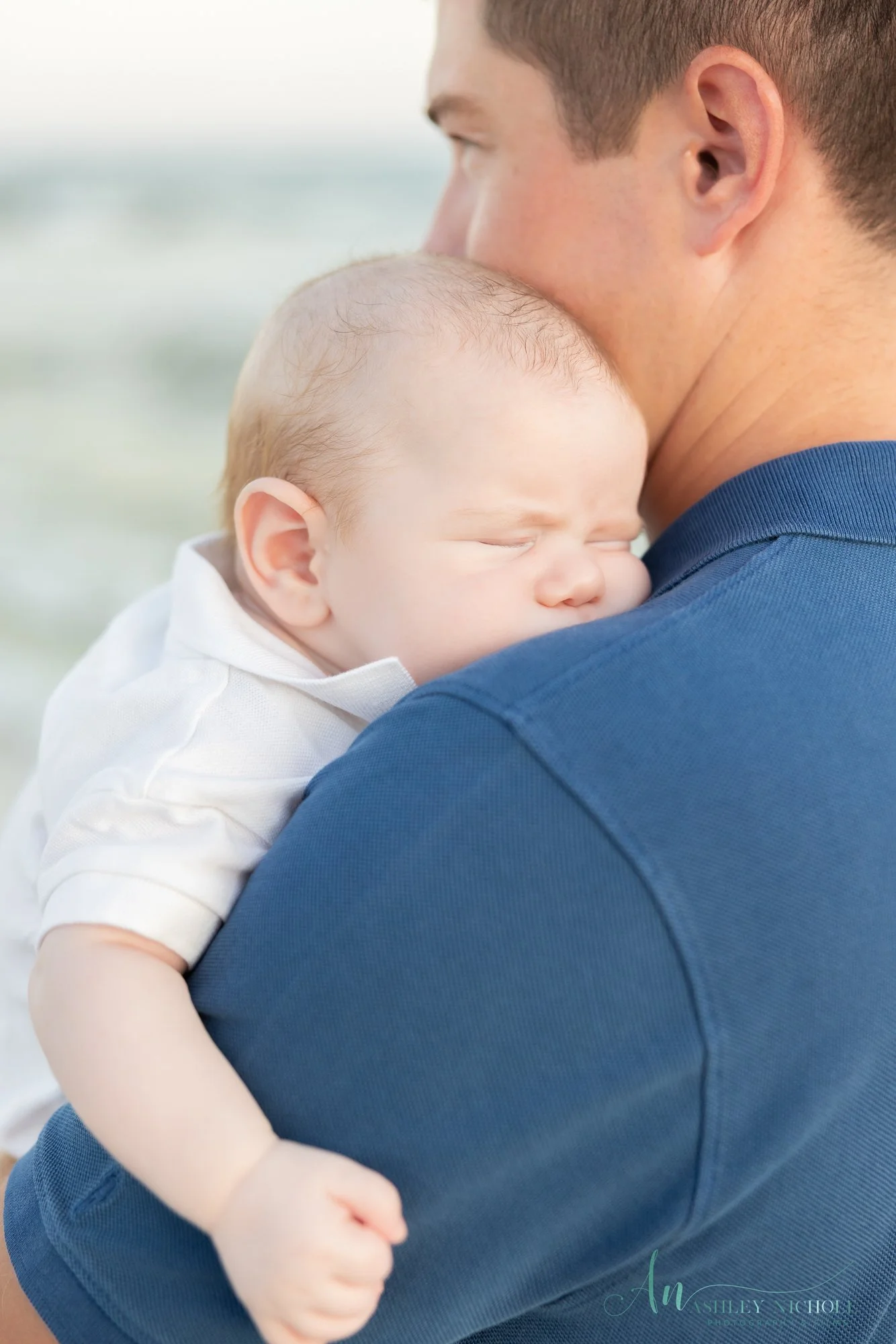 A man holding a sleeping baby close to his chest, outdoors near a beach or water.