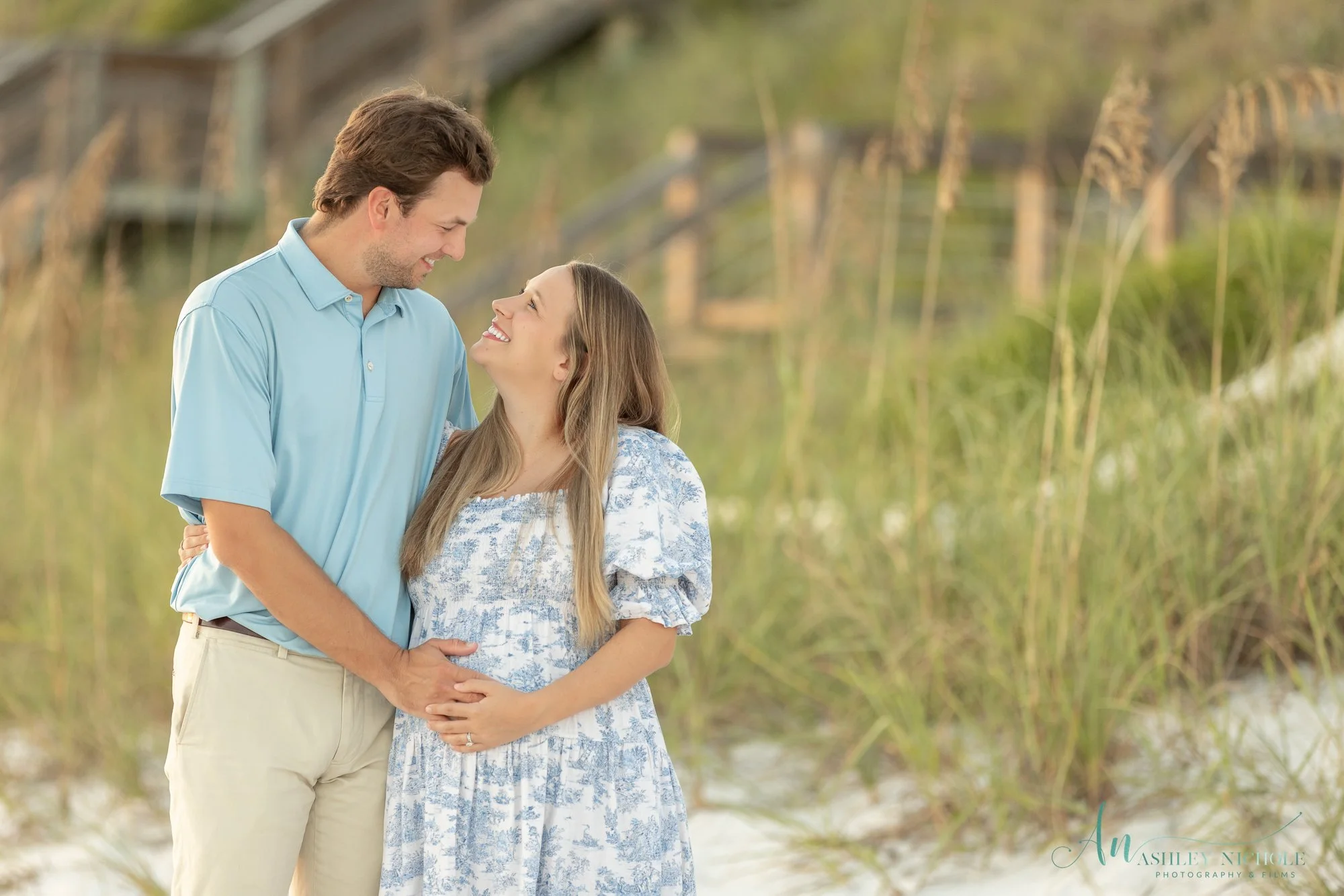 A smiling couple standing close together on a beach, the man with his arm around the woman, holding her belly as if pregnant, with a wooden bridge and tall grasses in the background.
