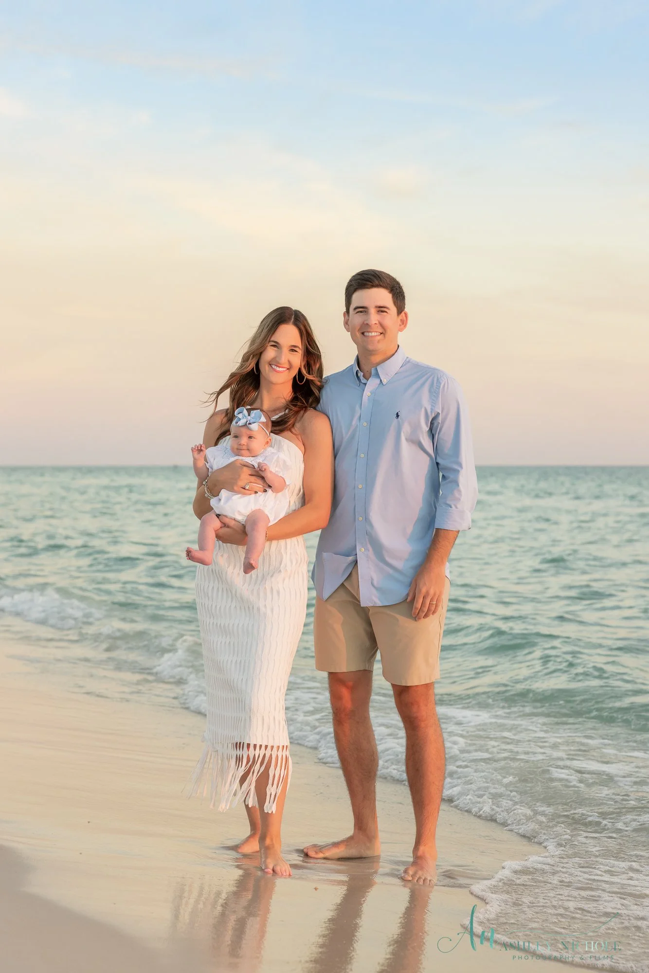 A family standing barefoot on a beach at sunset, with the ocean in the background. The woman holds a baby girl, and a man stands beside her, all smiling.