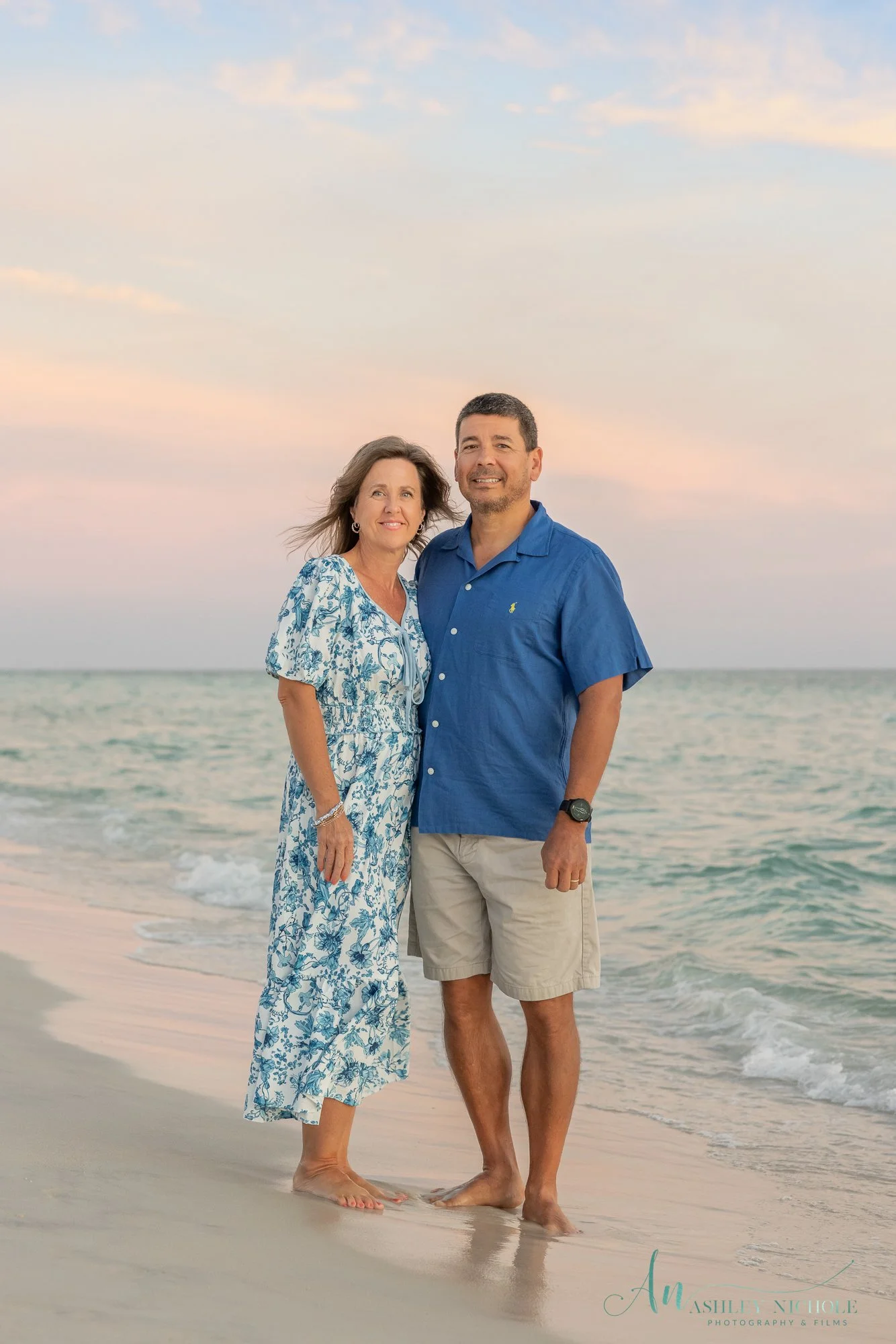 A middle-aged couple stands barefoot on the beach at sunset, smiling at the camera. The woman wears a blue and white floral dress, and the man wears a blue short-sleeve shirt and khaki shorts. The ocean and a pastel-colored sky are in the background.