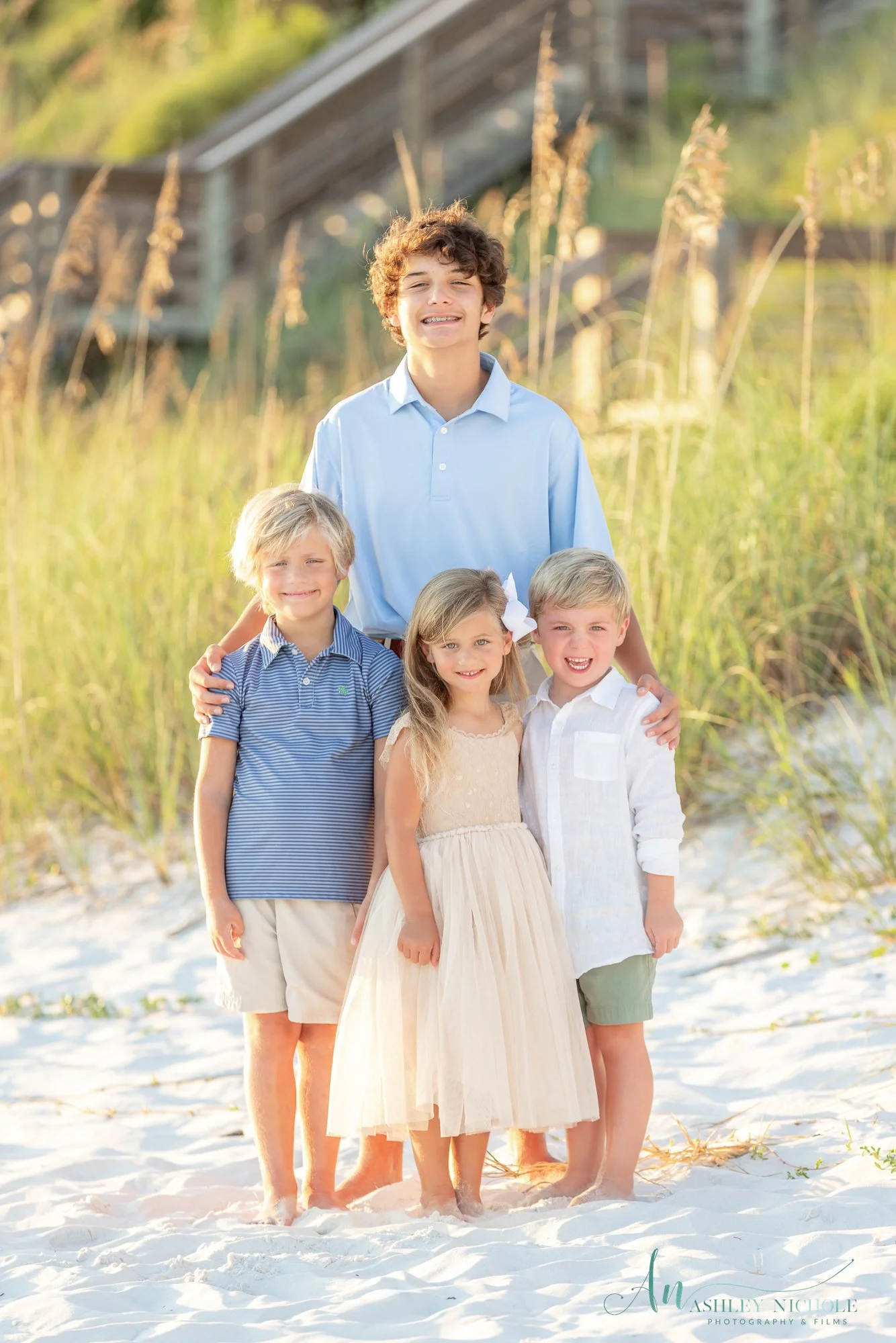 A teenage boy and four young children standing on a sandy beach with grass and a wooden pier in the background, smiling at the camera.