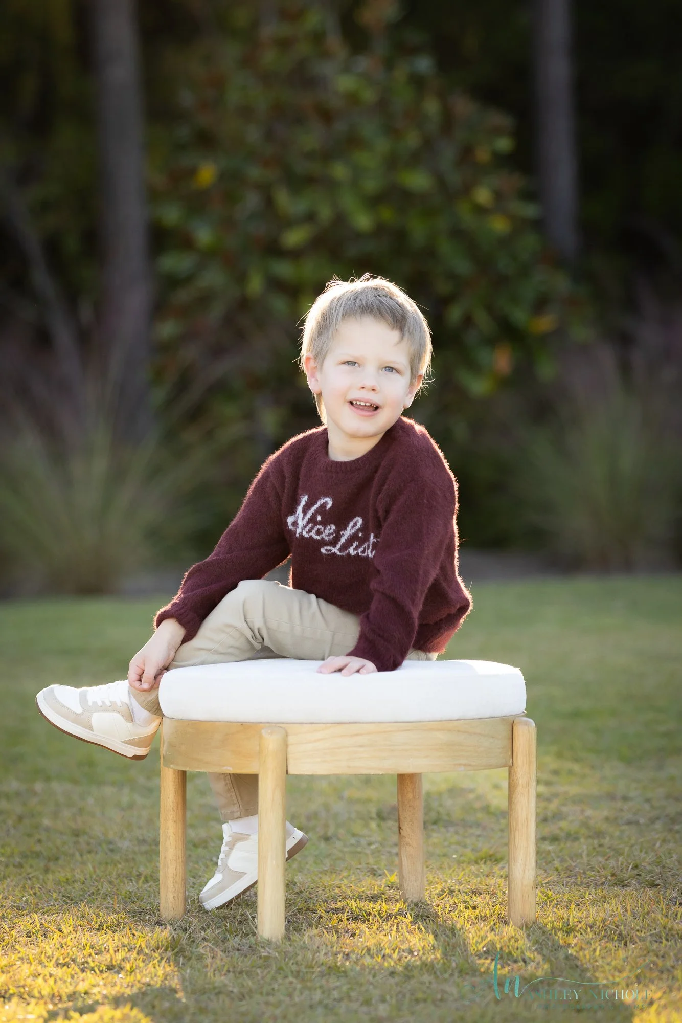 A young boy sitting on a white cushion on a wooden bench outdoors, wearing a maroon sweater and beige pants, with a puzzled expression. The background features trees and greenery.