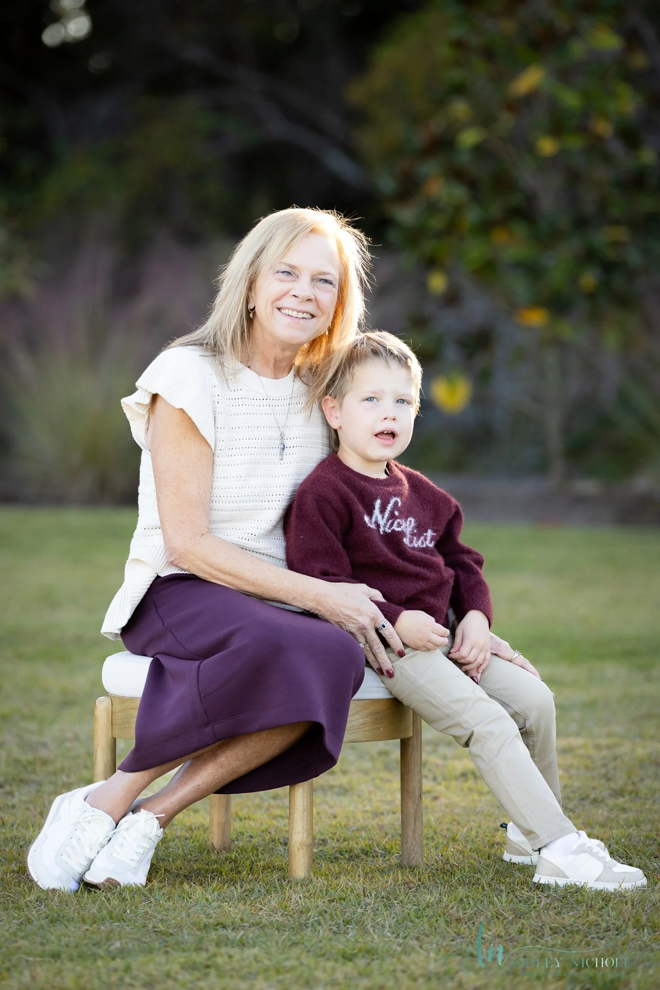 An elderly woman and a young boy sitting outdoors on a small bench, smiling at the camera. The woman has blonde hair and is wearing a white top with a purple skirt, while the boy has light brown hair and is wearing a maroon sweater and beige pants.