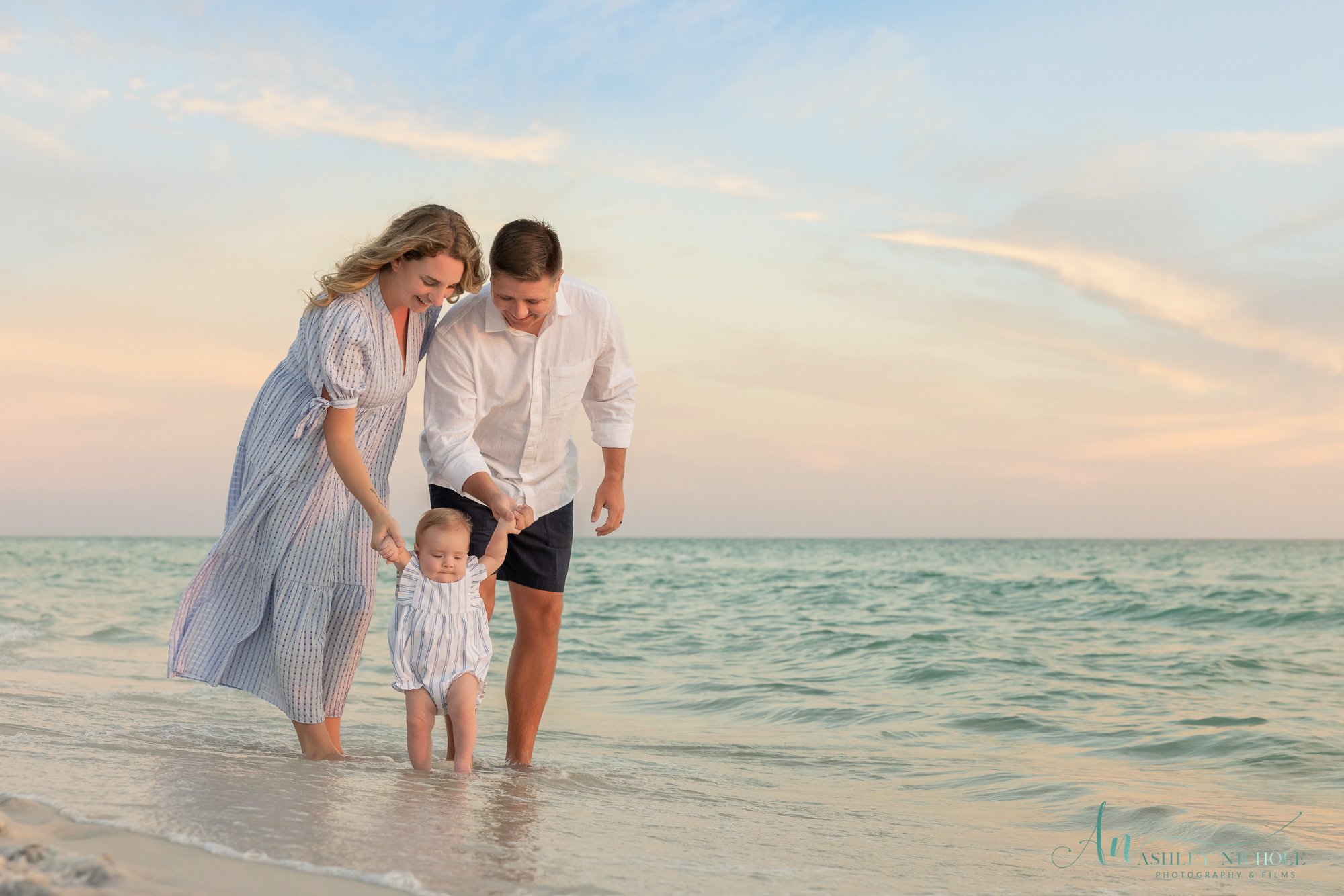 A family of three, a woman, a man, and a young child, walking hand in hand along the shoreline at the beach during sunset.