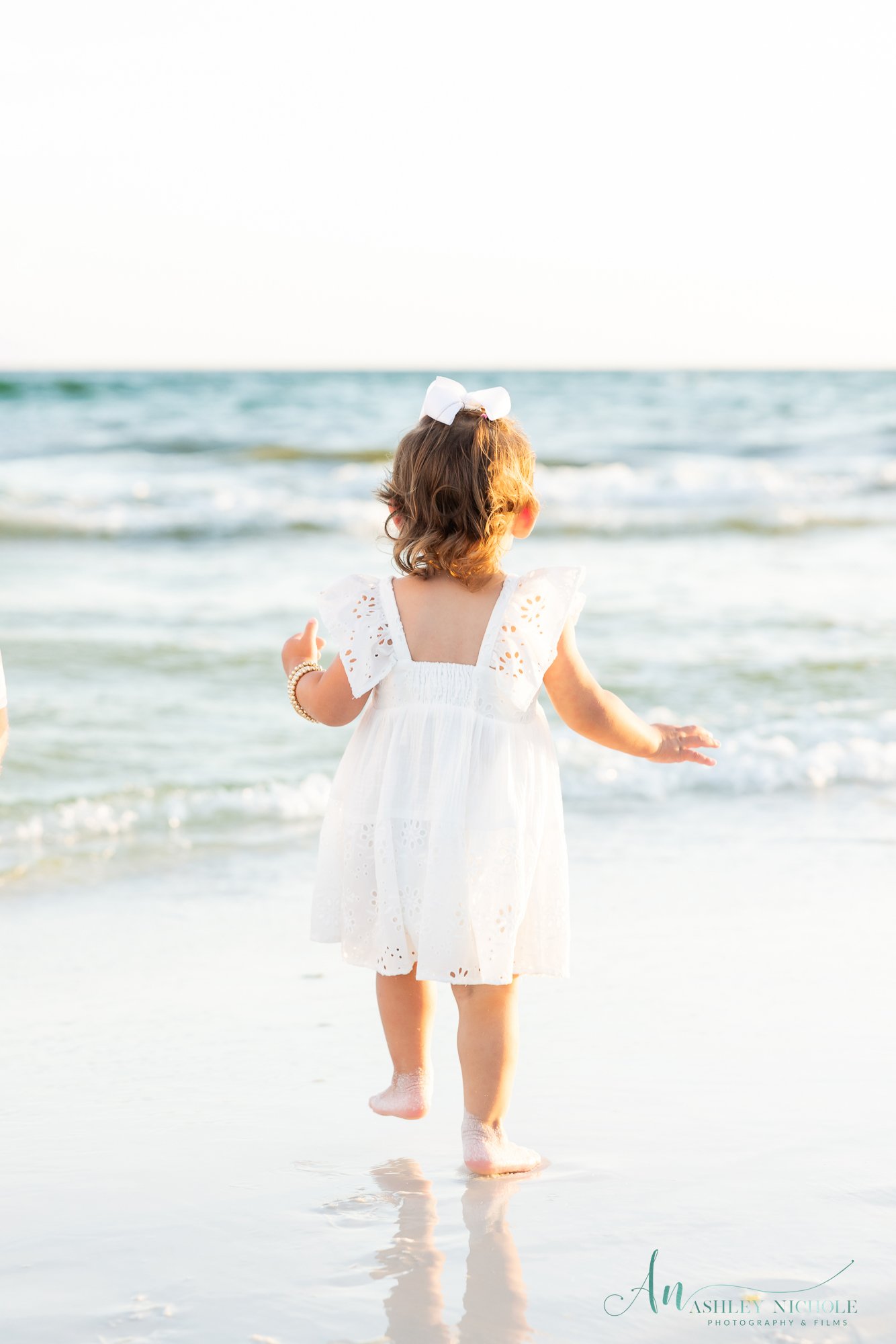 Young girl in a white dress with eyelet details, walking barefoot on the beach towards the ocean, with her back facing the camera, wearing a white bow in her hair.