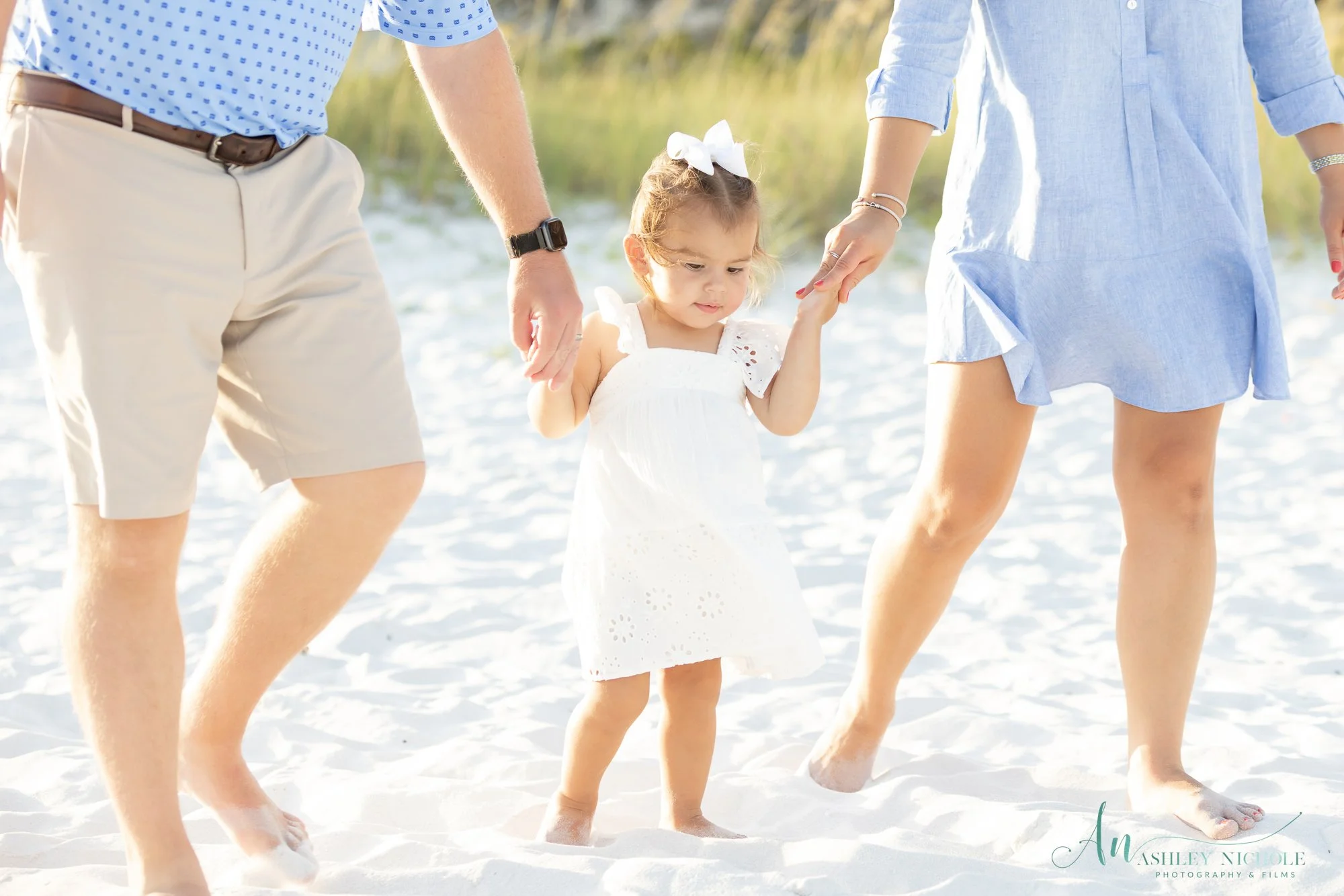 A young girl in a white dress holding hands with two adults, walking on a sandy beach.
