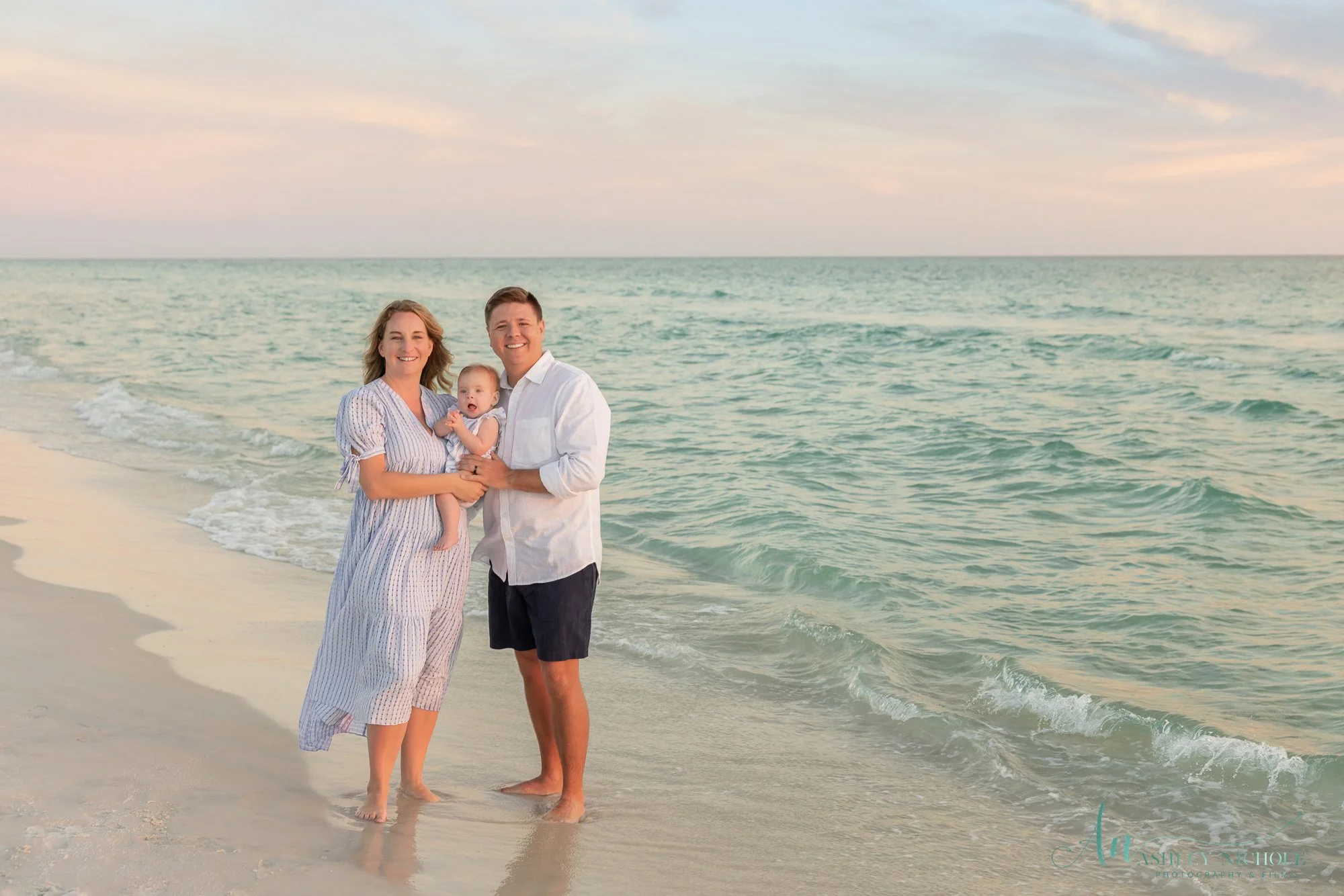 A family of three standing on the beach, holding each other, with ocean waves and a pastel-colored sky in the background.
