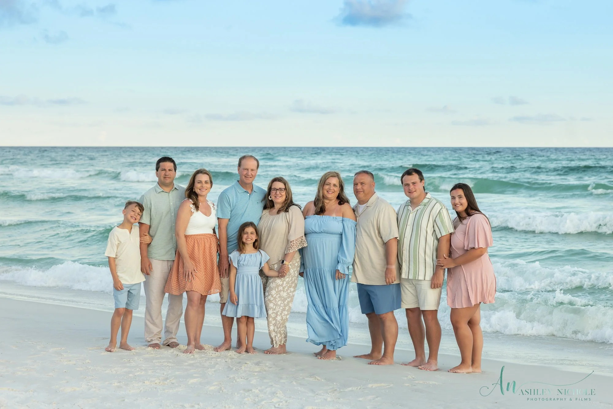 Family of eleven standing on a beach with ocean waves in the background, dressed in beachwear and smiling.