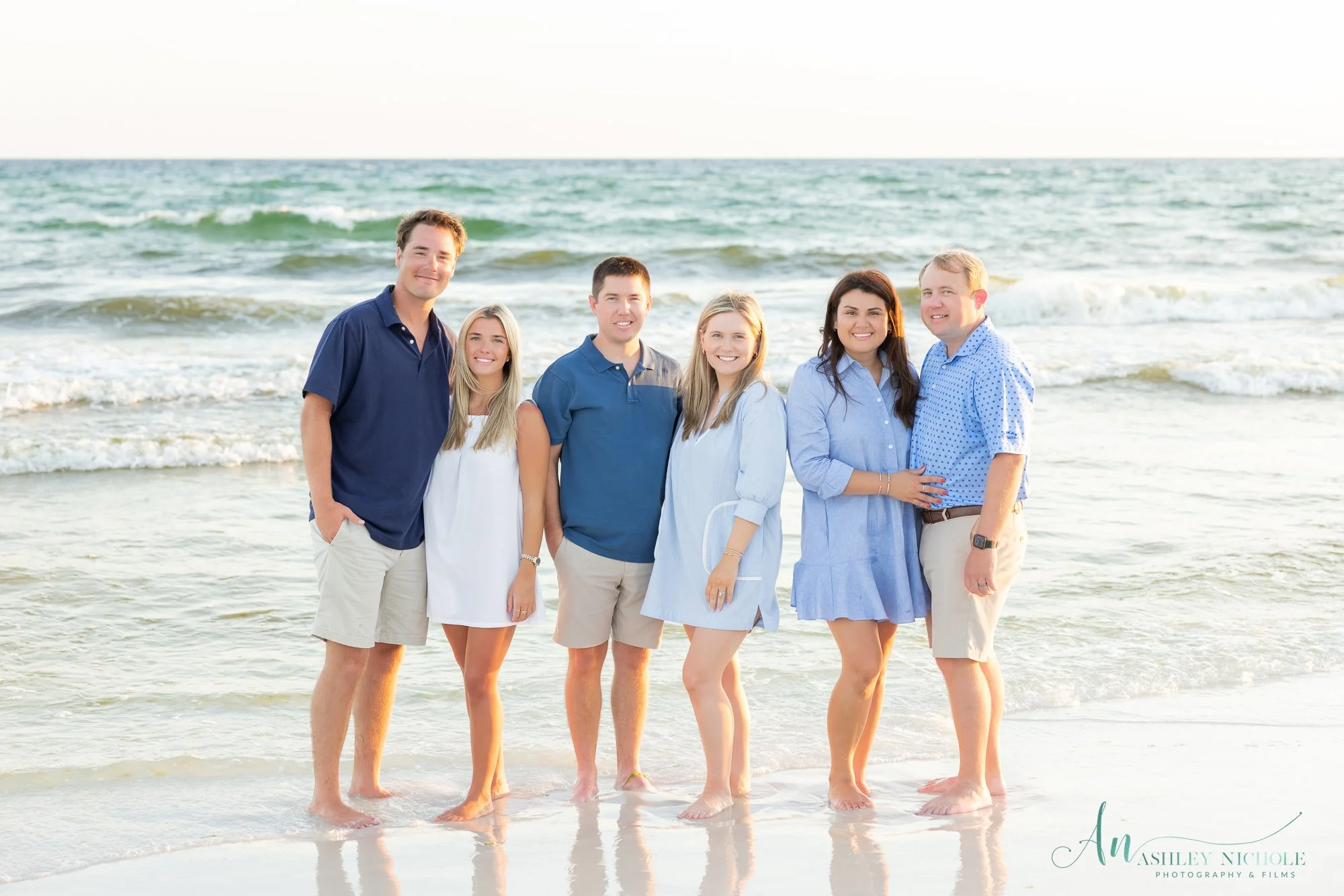 Group of six friends standing on the beach near the water with ocean waves behind them, smiling and dressed in casual summer outfits.