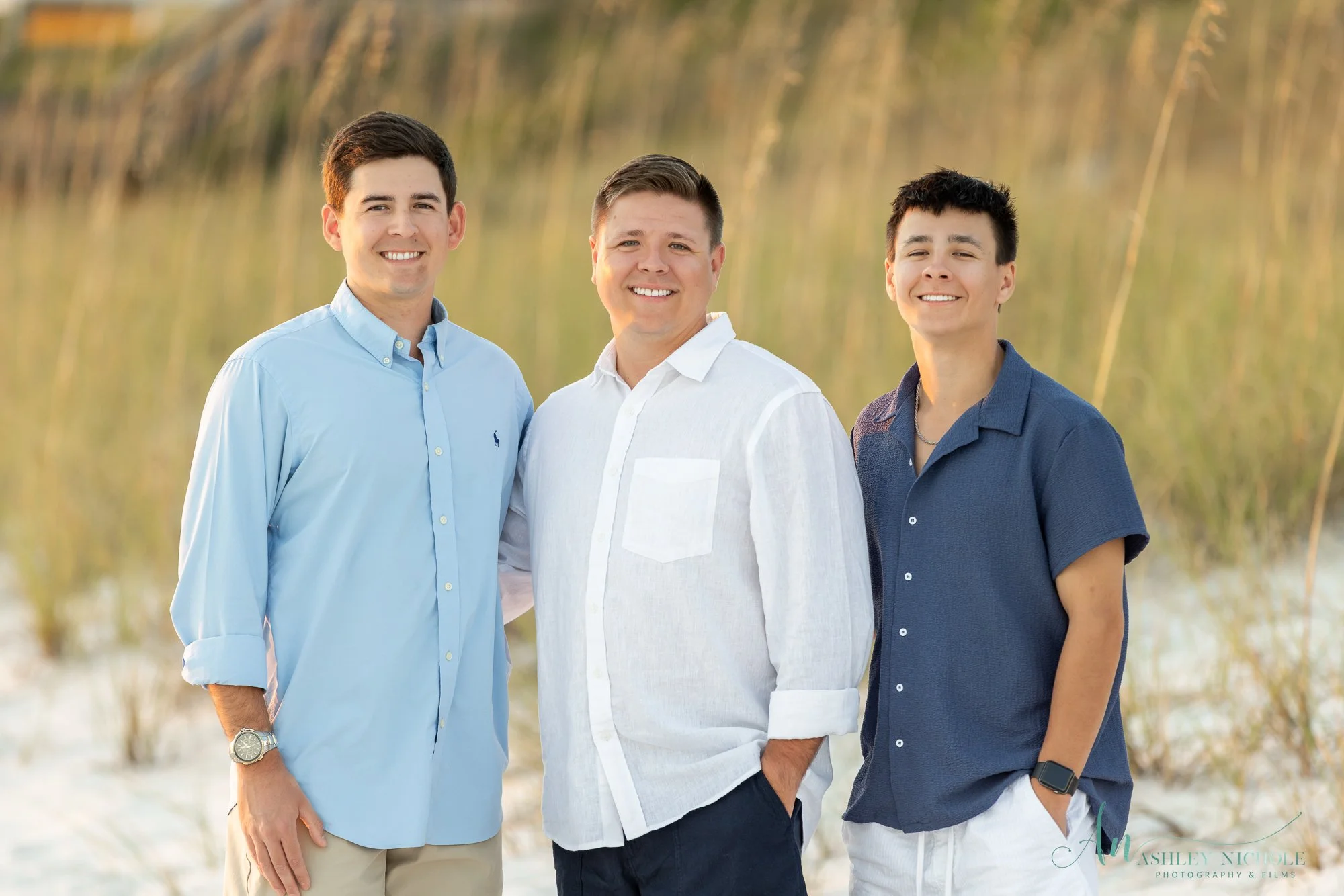 Three smiling men stand together outdoors on a sandy area with grass in the background.