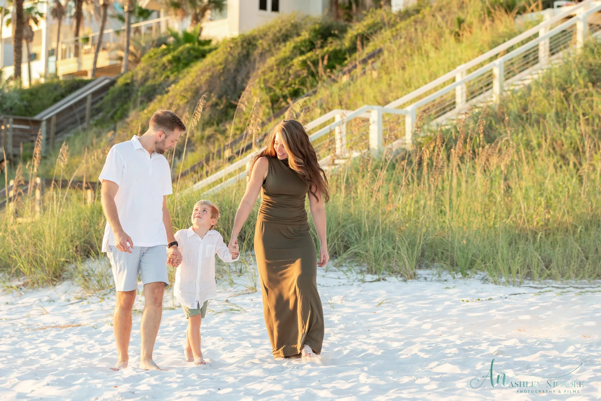 A family of three walking barefoot on the beach, holding hands and smiling, with sand dunes and grass in the background during sunset.