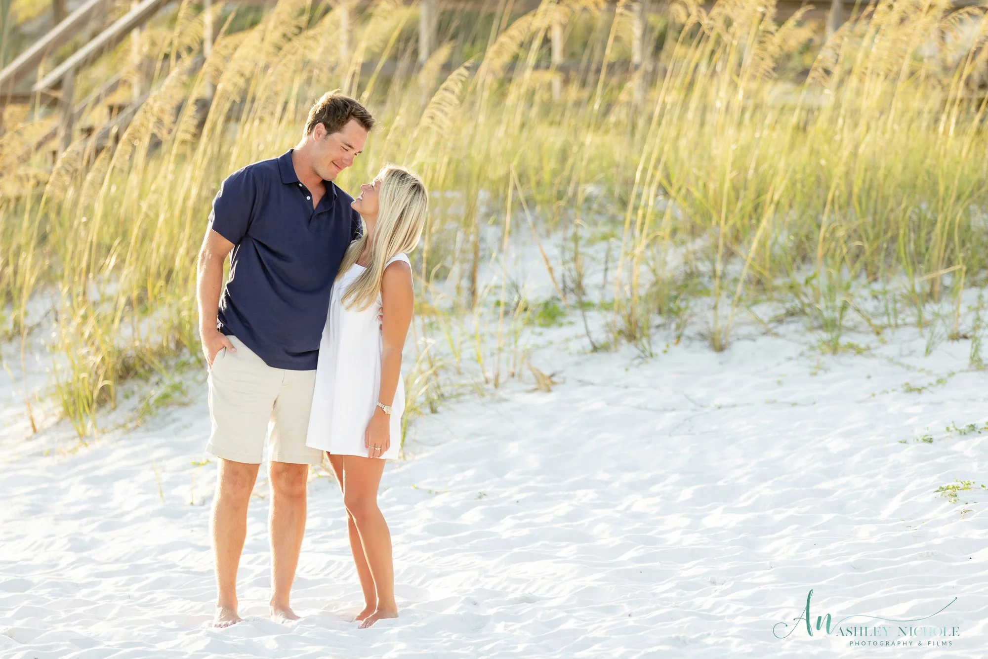 A man and woman standing on a sandy beach, smiling and looking at each other, with tall grass and a wooden structure in the background.