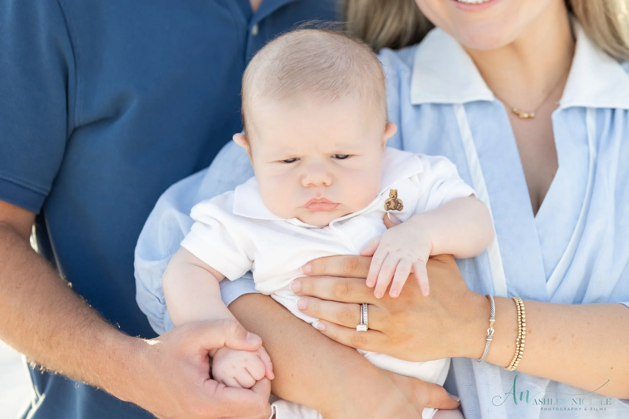 Close-up of a baby being held by a woman, with a man also holding the baby's hand. The baby has a serious expression and is wearing a white shirt with a small bear embroidered on it.