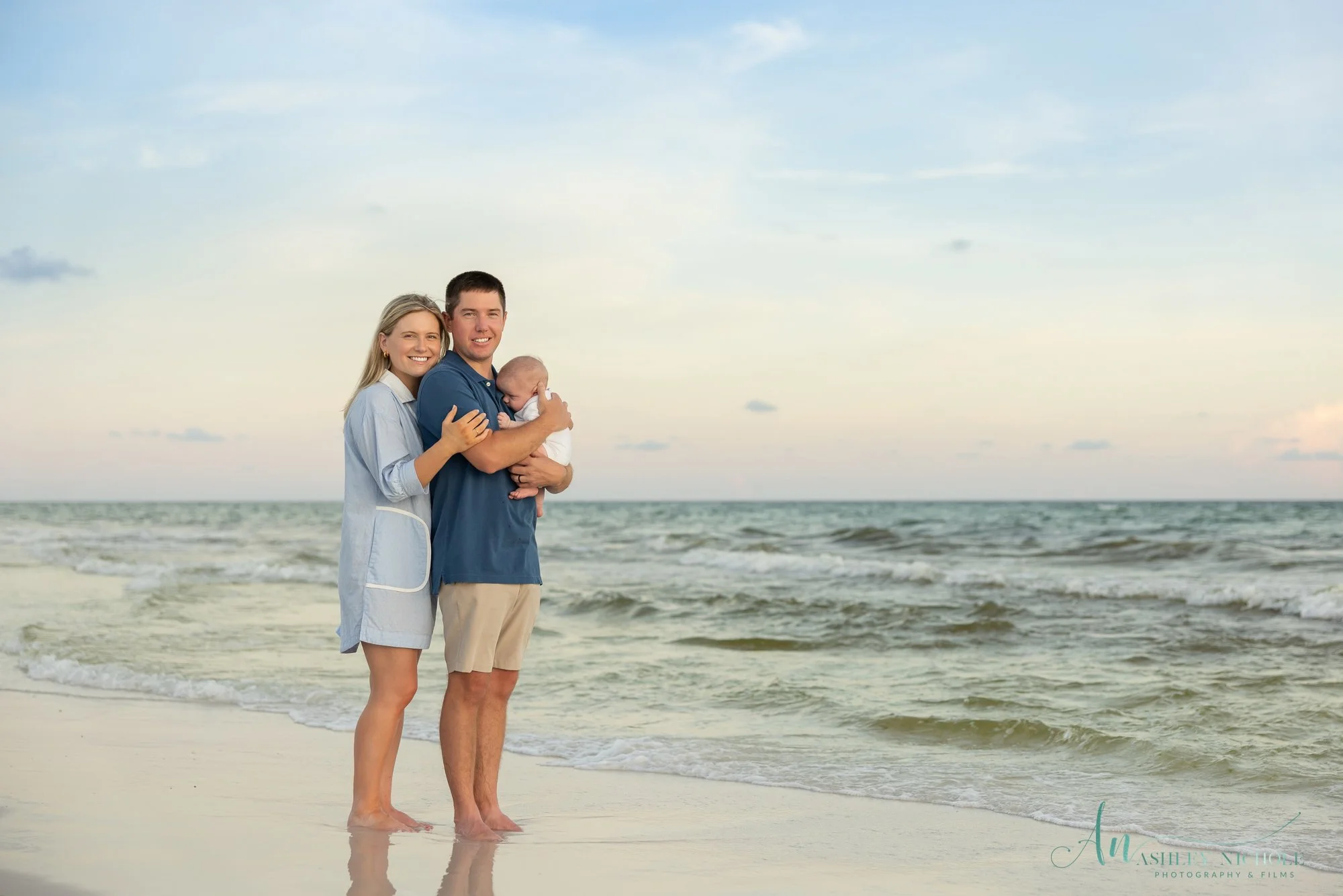 A happy family of three, a mother, father, and their baby, standing on the beach with the ocean in the background during sunset, smiling at the camera.
