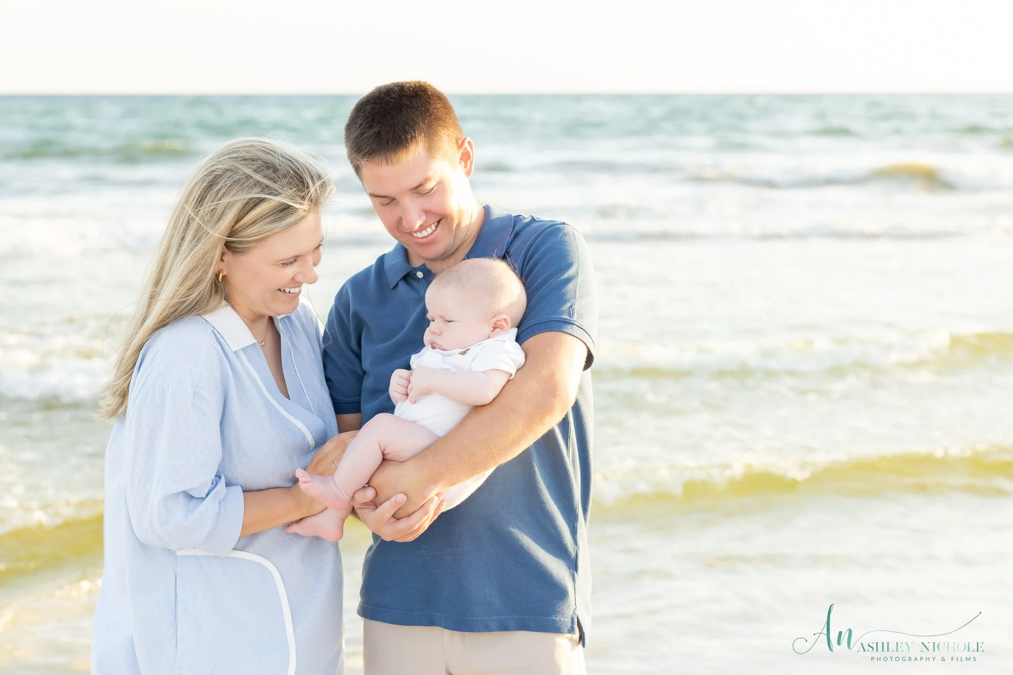 A happy family of three standing on the beach, a woman and a man holding a baby.